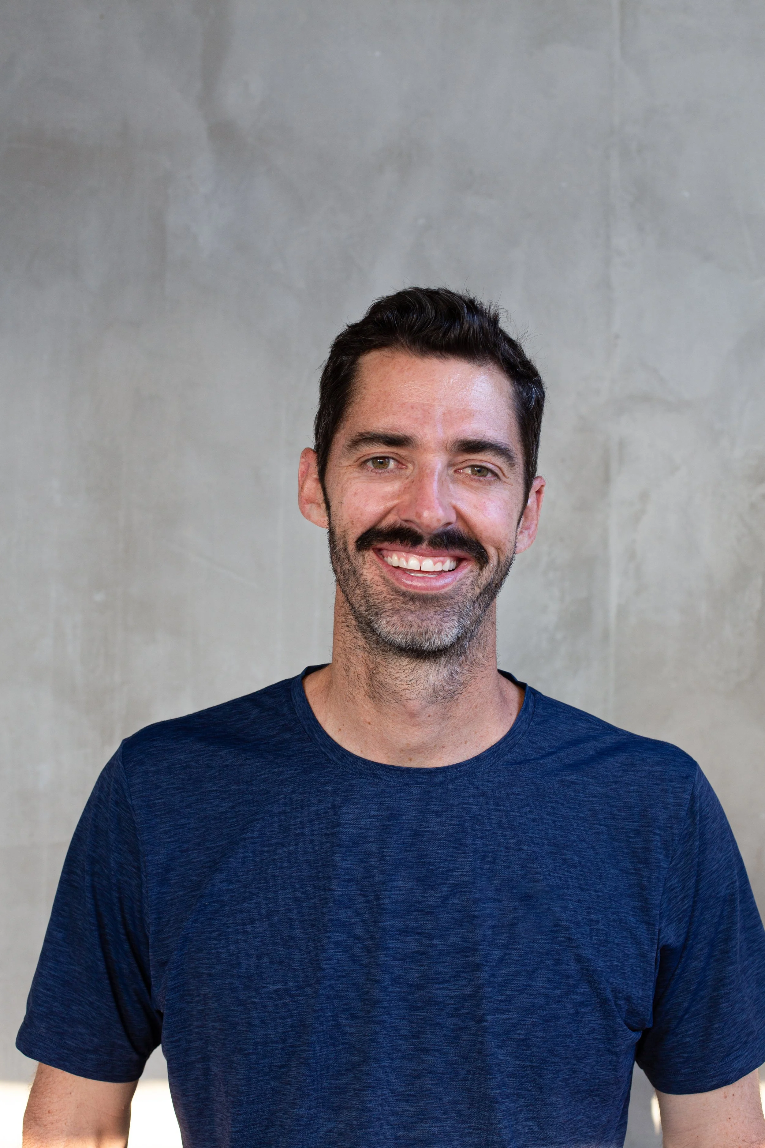 A man with dark hair and a beard, smiling, wearing a navy blue t-shirt, standing against a gray concrete wall.