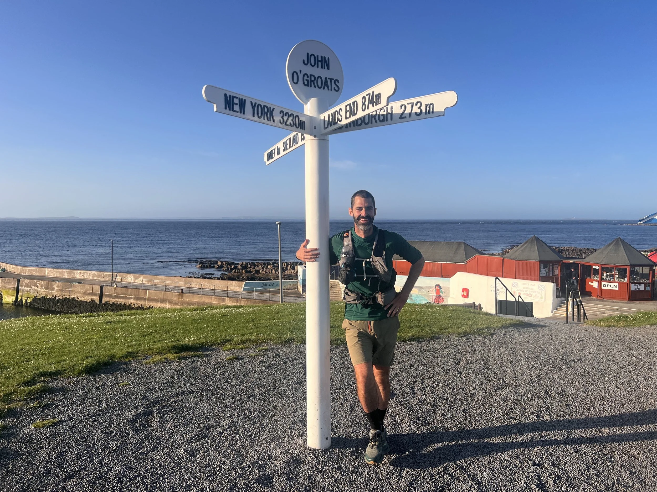 A man in outdoor clothing standing next to a signpost on a gravel path by the ocean. The signpost has multiple directions including New York, Edinburgh, and the North Sea. The ocean is visible in the background with a clear blue sky.