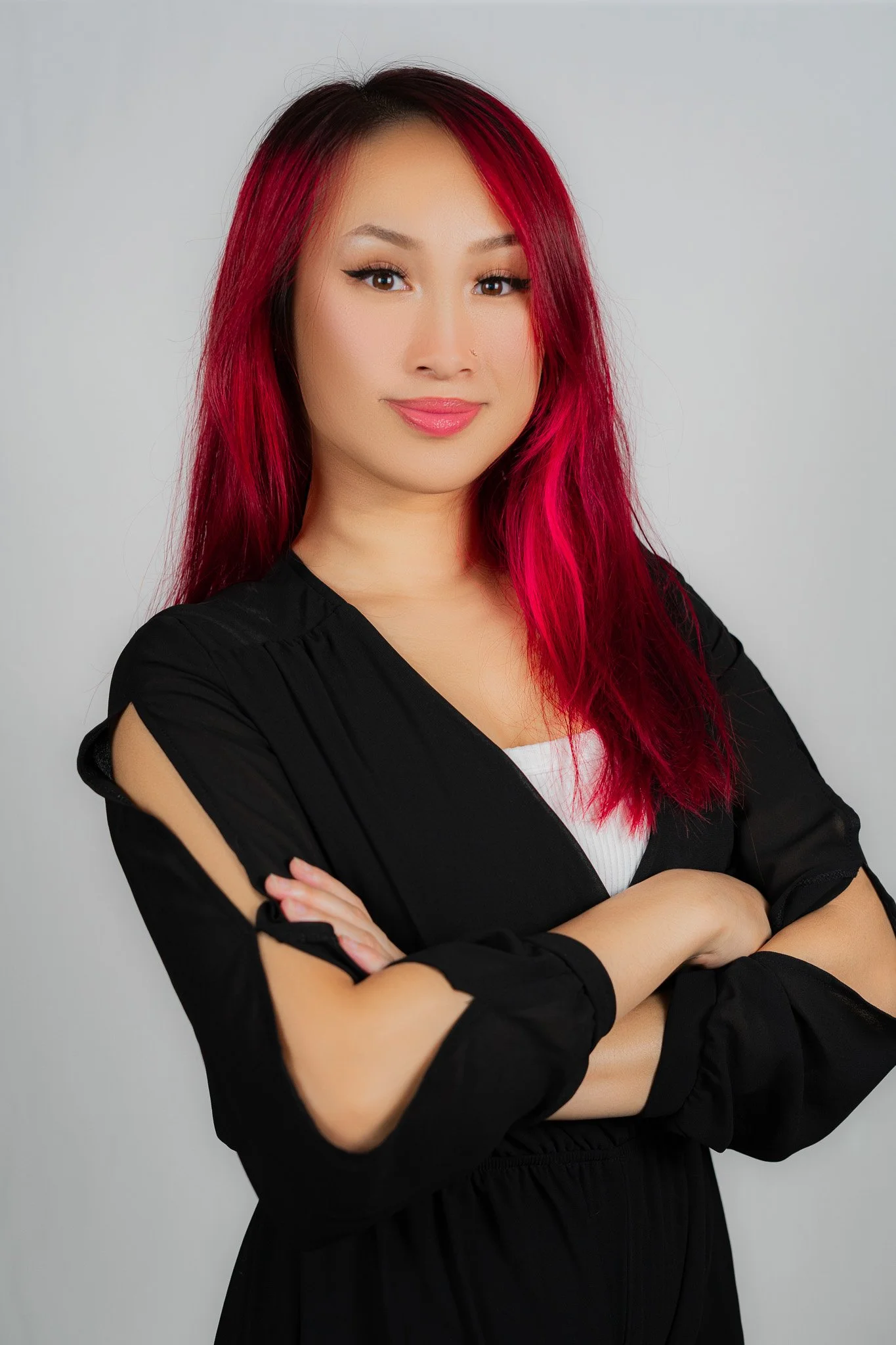 A woman with long, bright pink hair, wearing a black jacket with rolled-up sleeves and a white top, standing with arms crossed and smiling slightly.