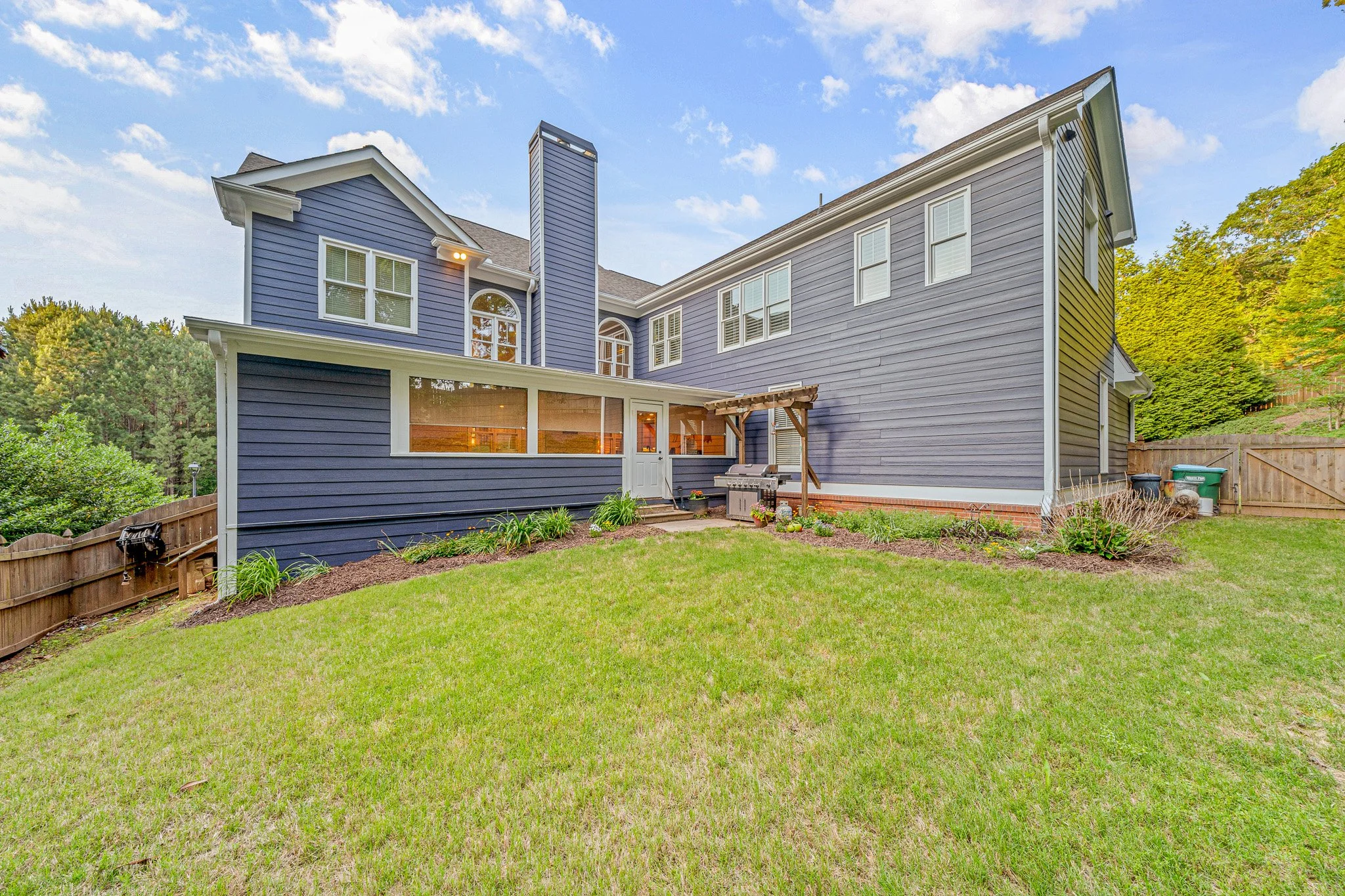 Backyard view of a blue two-story house with a grassy lawn, a fenced yard, and a small patio area with a grill and garden decorations.