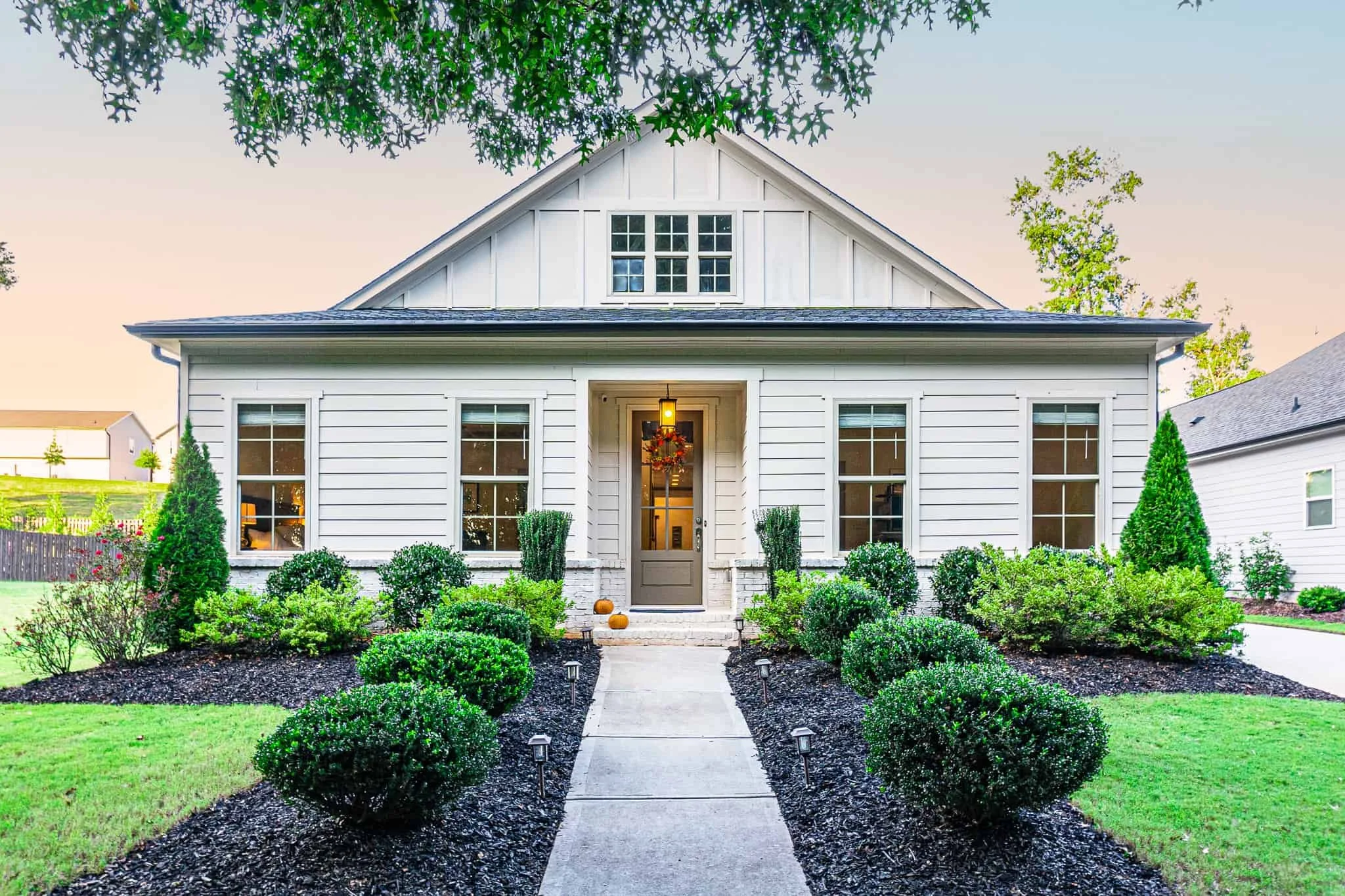 Front view of a white house with a peaked roof, four large windows, and a central front door decorated for fall. Well-manicured shrubs and bushes line the walkway leading to the porch, surrounded by dark mulch and small outdoor lights.