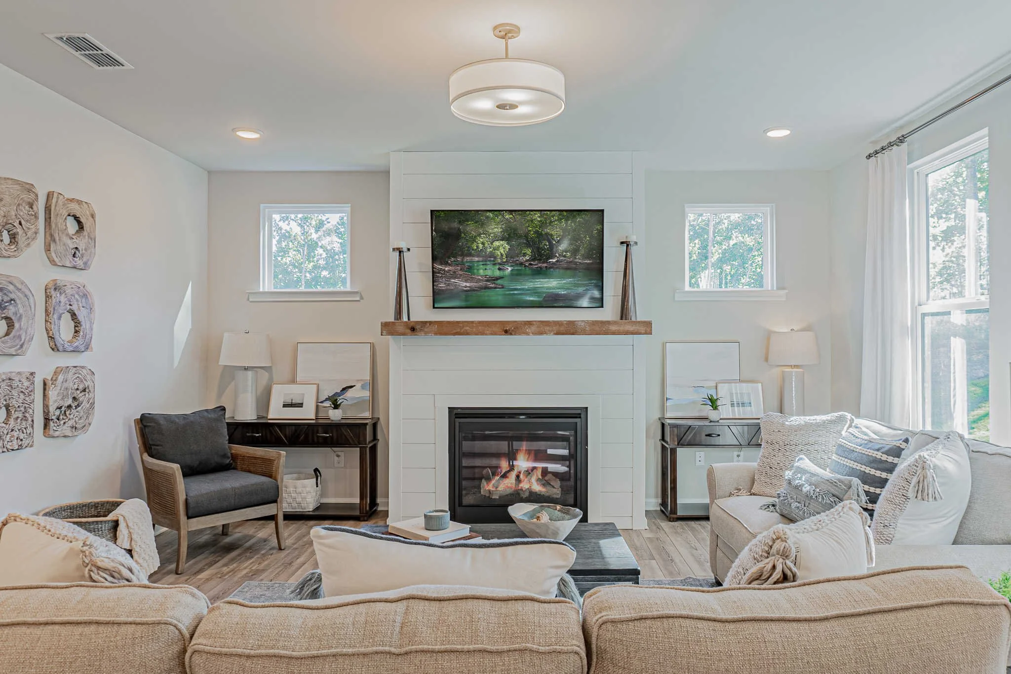 Living room with white walls, a white fireplace, and a flat-screen TV mounted above the fireplace. There are two small windows, one on each side of the TV. The living room has a beige sofa with decorative pillows, a black side table with a lamp on each side of the fireplace, and a gray armchair on the left. There are artwork pieces on the wall to the left and a wooden mantel above the fireplace.
