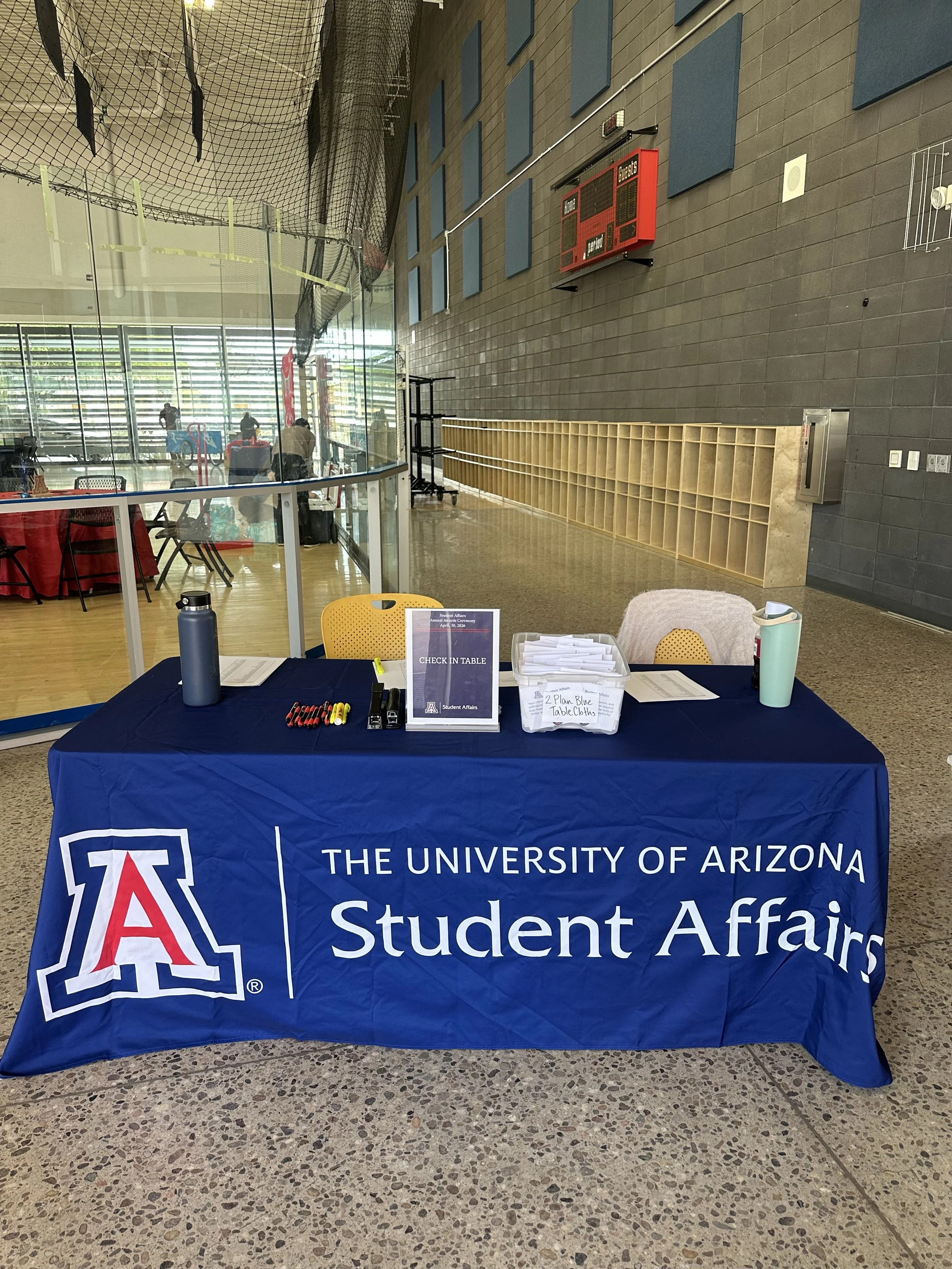 A table with a blue tablecloth displaying the university's logo and name, "The University of Arizona Student Affairs," along with a sign that says "Check-In Table," pens, and papers in an indoor facility with a netted area and blue wall panels in the