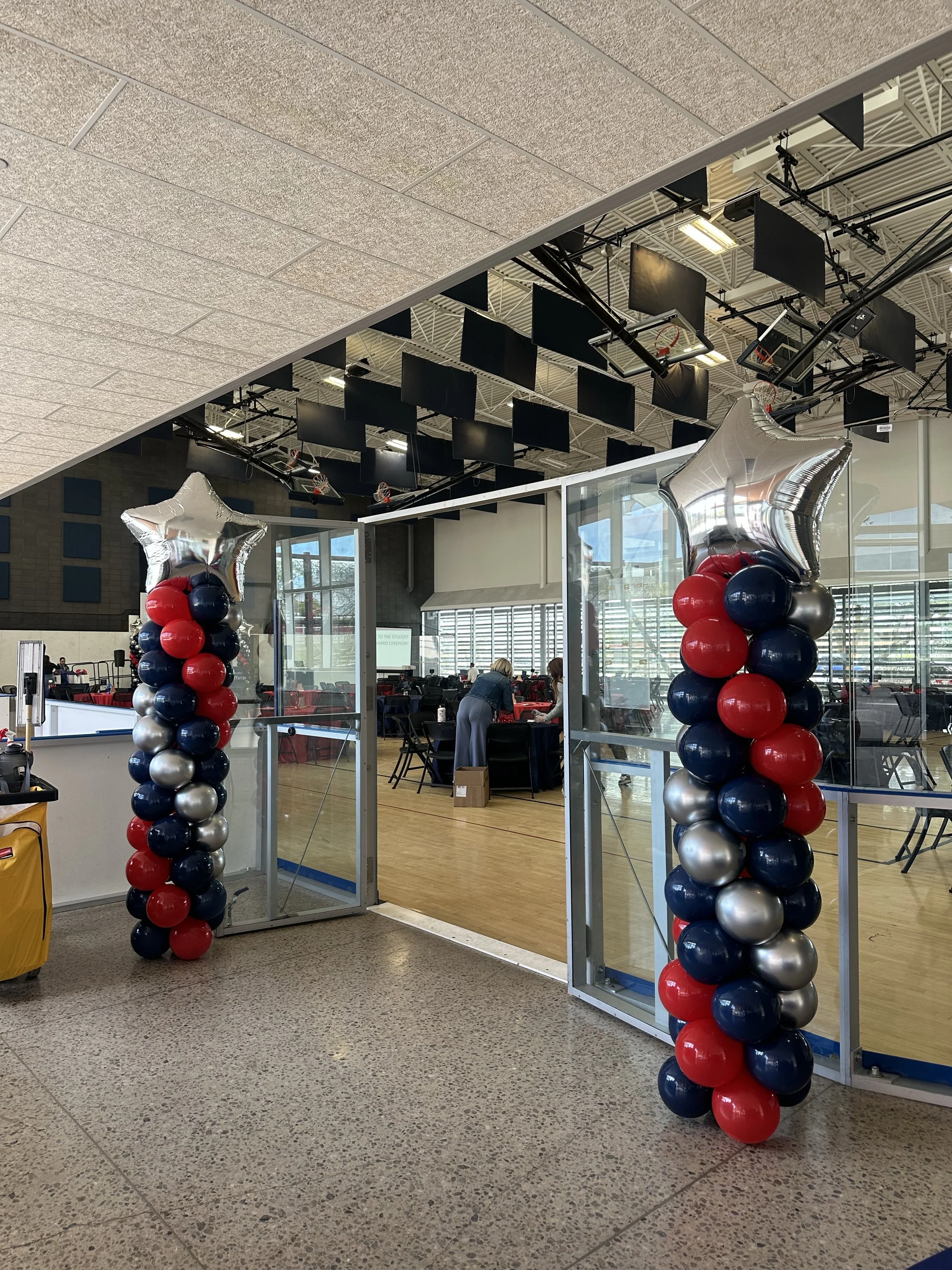 Decorative balloon pillars with star-shaped balloons at entrance of indoor event space, with tables and people visible inside.