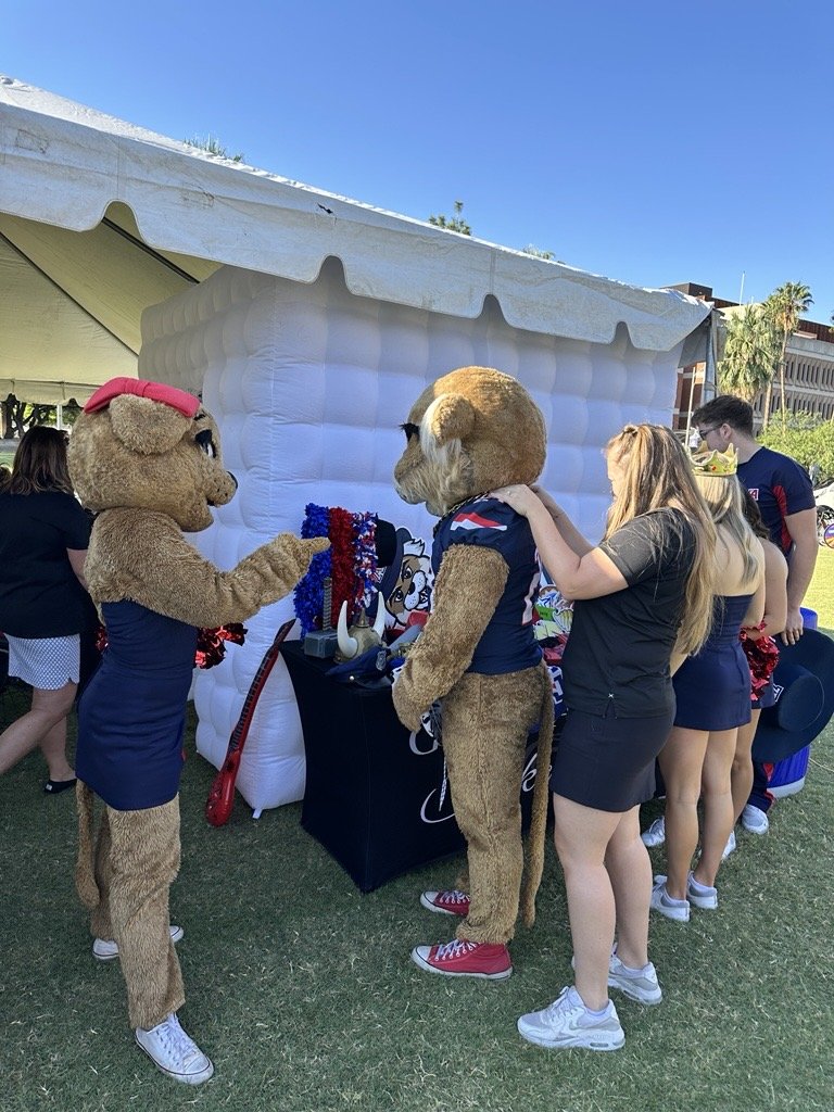 Two people in lion costumes are at an outdoor event, engaging with two women. One woman has her arm around a lion's neck, while the other woman is in line behind them. The background features a white inflatable structure with a tent roof and other at