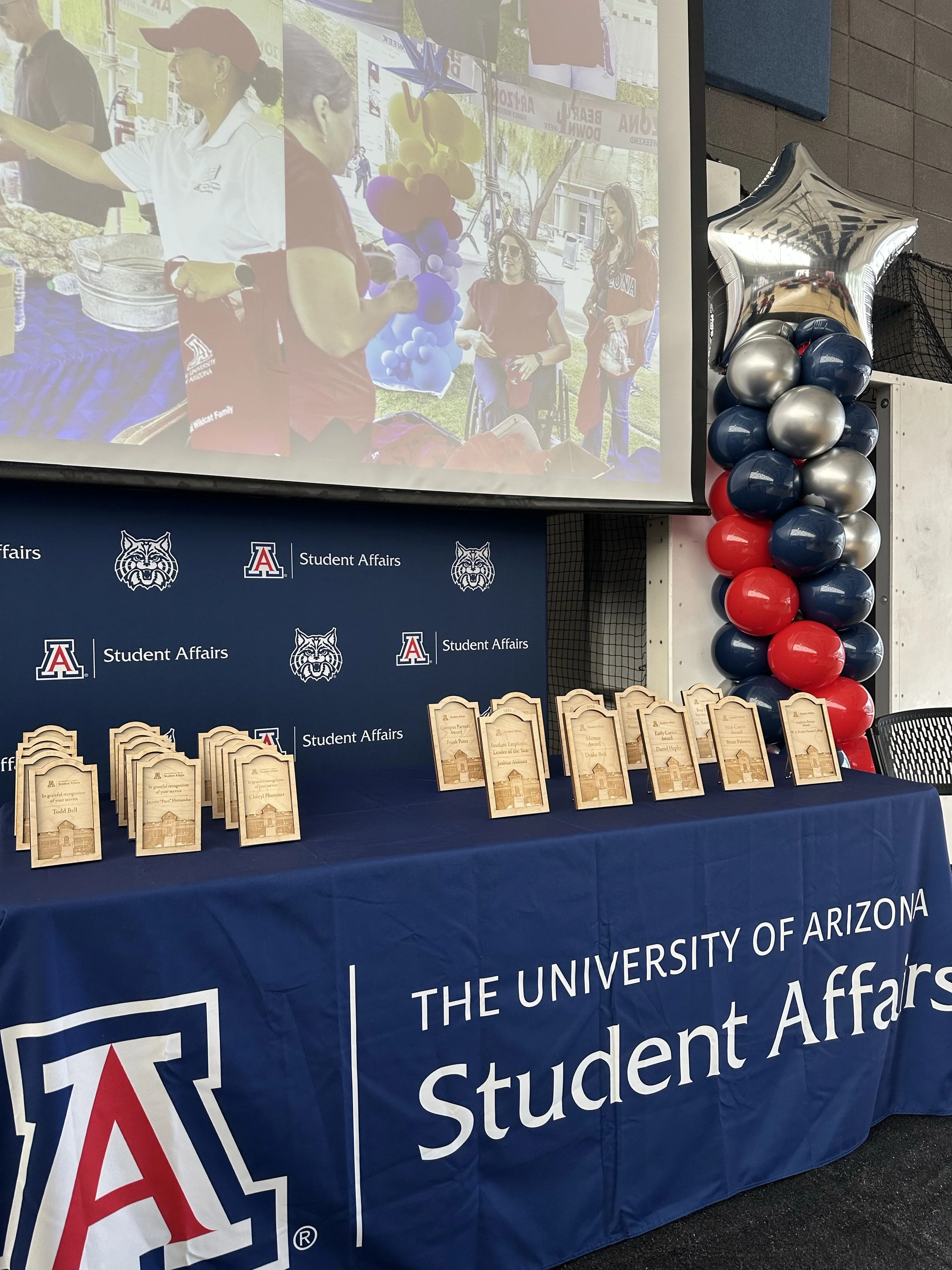 A table covered with a blue tablecloth displaying awards and plaques for the University of Arizona's Student Affairs department. Behind the table is a banner with the university's logo and branding, and on the right side, balloon decorations in red, 