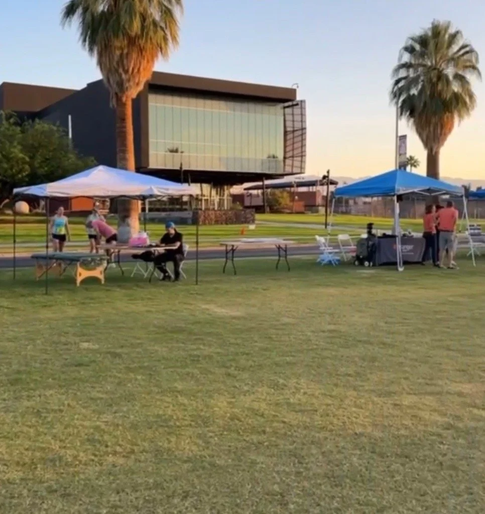 A group of people setting up tents and tables in an open grassy area with palm trees and a modern building in the background, during the evening.