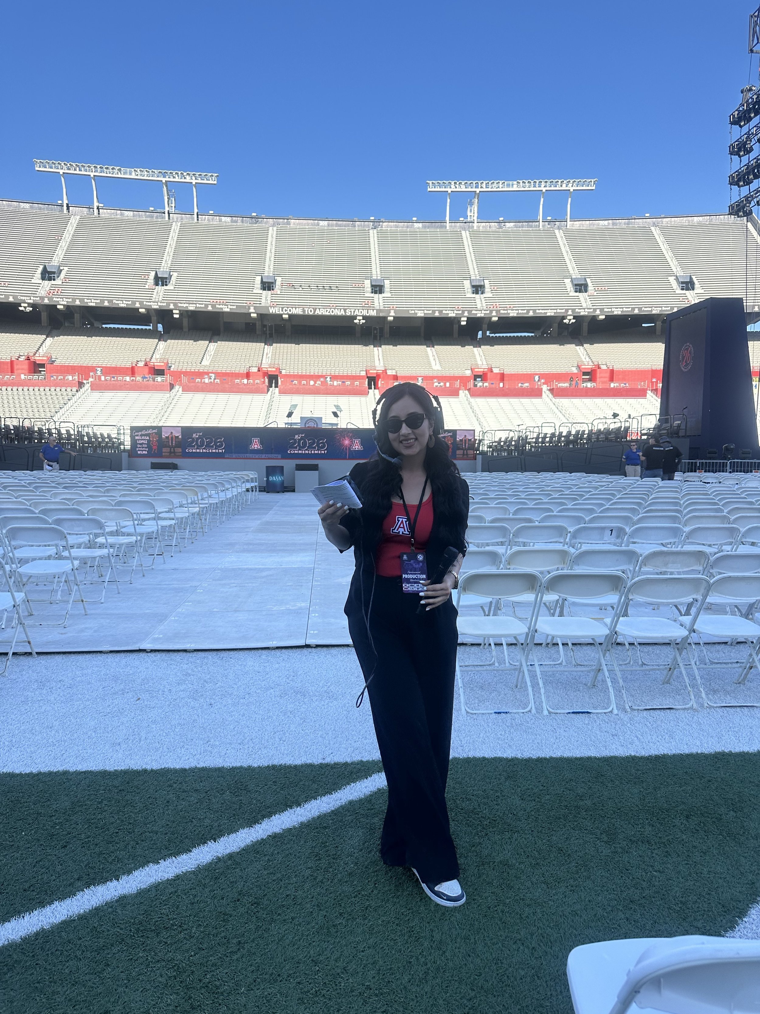 Woman wearing sunglasses and a headset standing on a football field, smiling, with empty stadium seats in the background, during daytime.