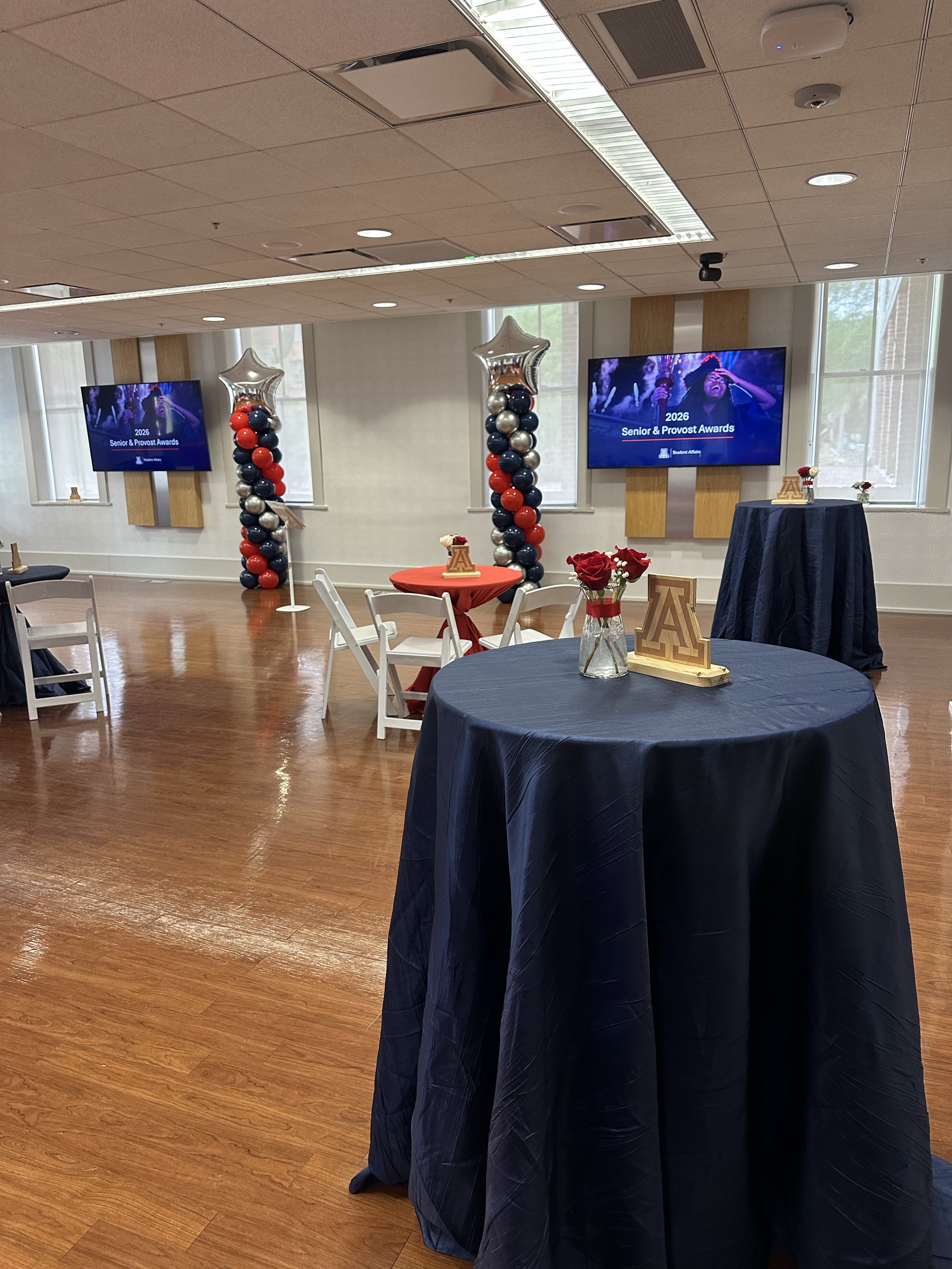 A decorated event room with round tables covered in navy tablecloths, featuring centerpieces with red roses. There are two large screens displaying a 2026 Senior & Provost Awards event, and balloon columns with star-shaped balloons and red, black, si