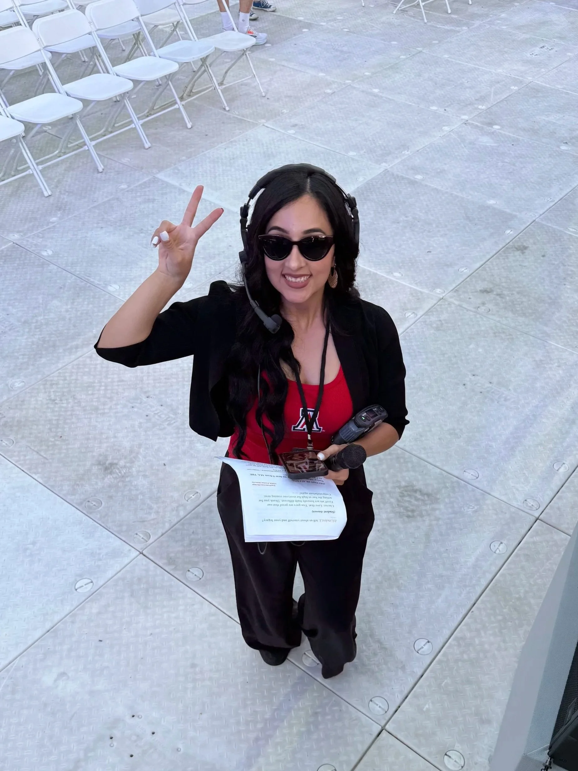 A woman with black hair, wearing sunglasses, a black jacket, and a red shirt, kneeling on a metal floor, making a peace sign with her right hand and holding a microphone, papers, and a walkie-talkie in her left hand.