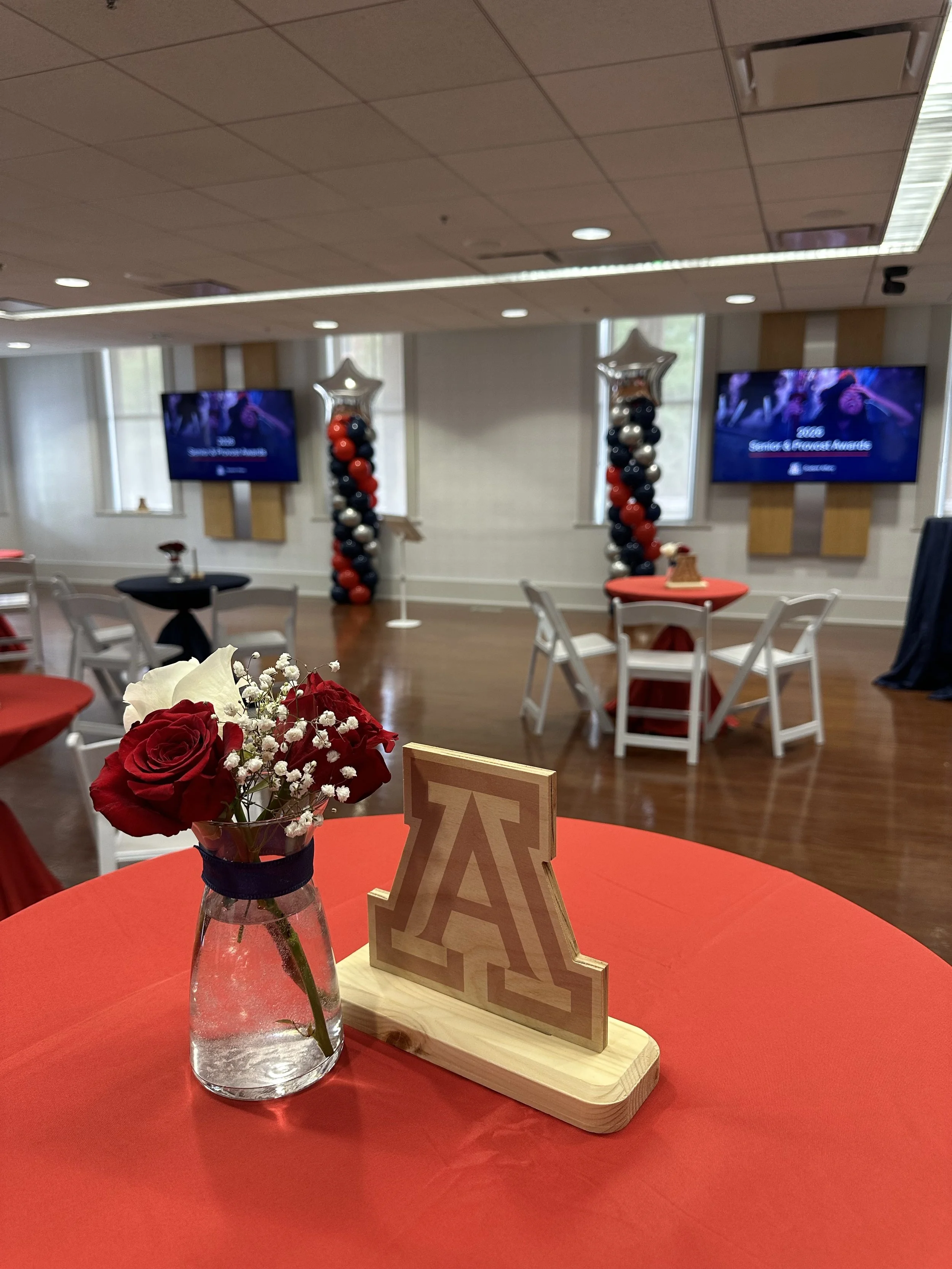 A red table with a glass vase containing red and white flowers, a wooden 'Alpha Tau' fraternity symbol, and a blurred background of a decorated room with television screens and balloon columns.