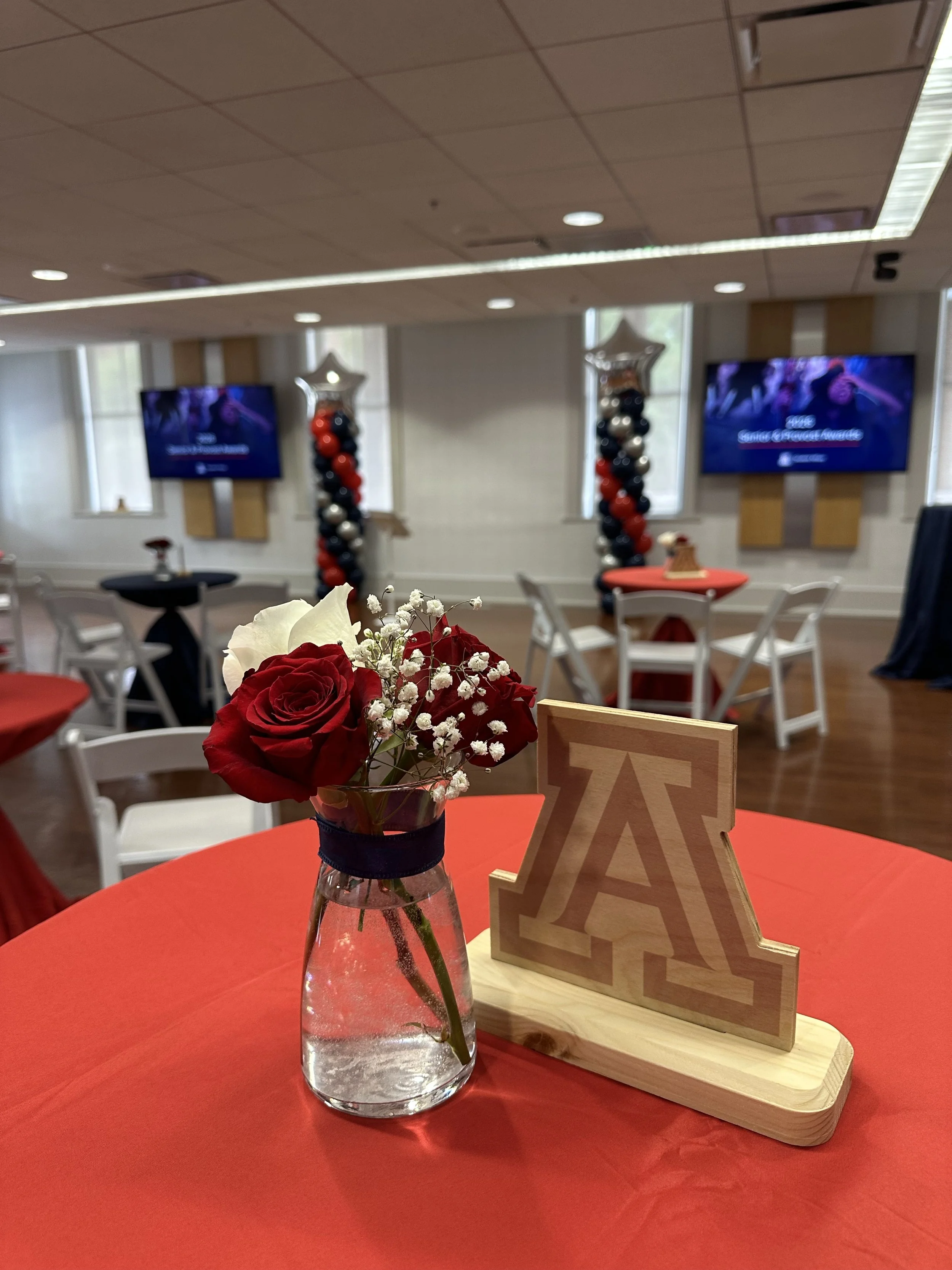 Centerpieces and decorations for a graduation celebration. A vase with red and white roses and baby's breath on a red table. A wooden 'A' letter and a wood-backed sign with a star-shaped balloon arch in the background.