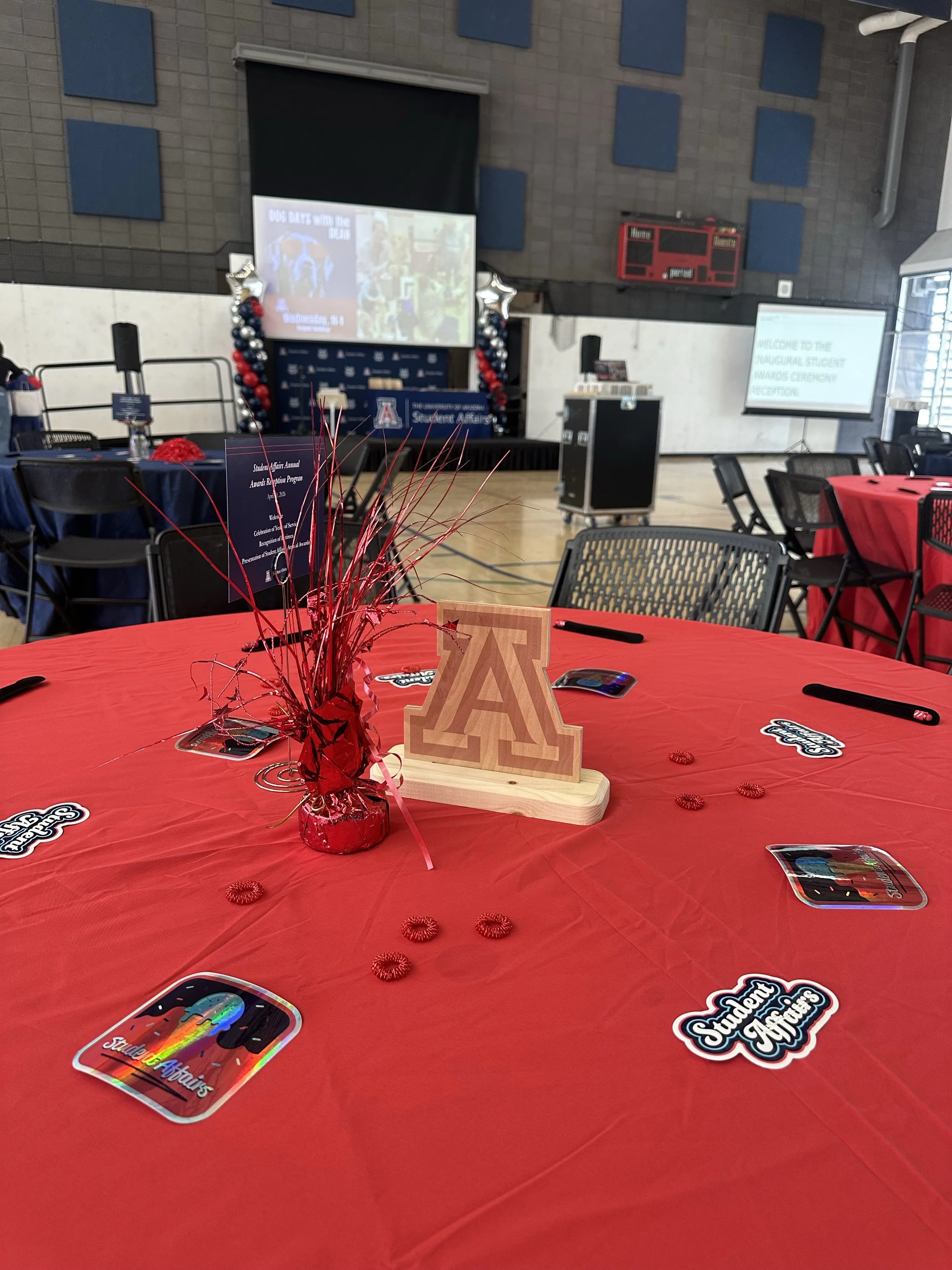 A decorated conference table with a red tablecloth, a wooden 'A' letter, rainbow-colored Student Affairs sticker, a rainbow-colored sticker, small red pom-poms, and red metallic decorative grass in a vase serving as a centerpiece. In the background, 