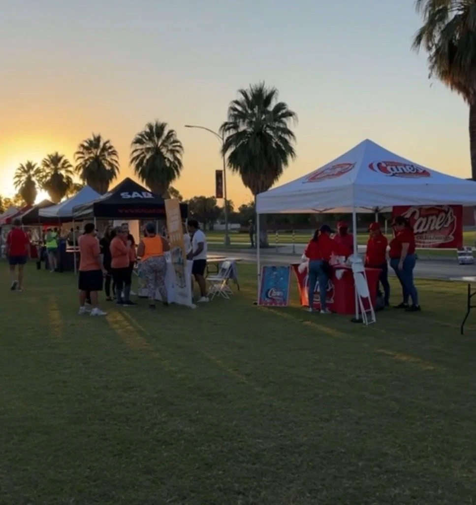 A row of food and promotional tents set up outdoors at sunset, with people walking and gathering around, palm trees in the background, and a grassy area in the foreground.