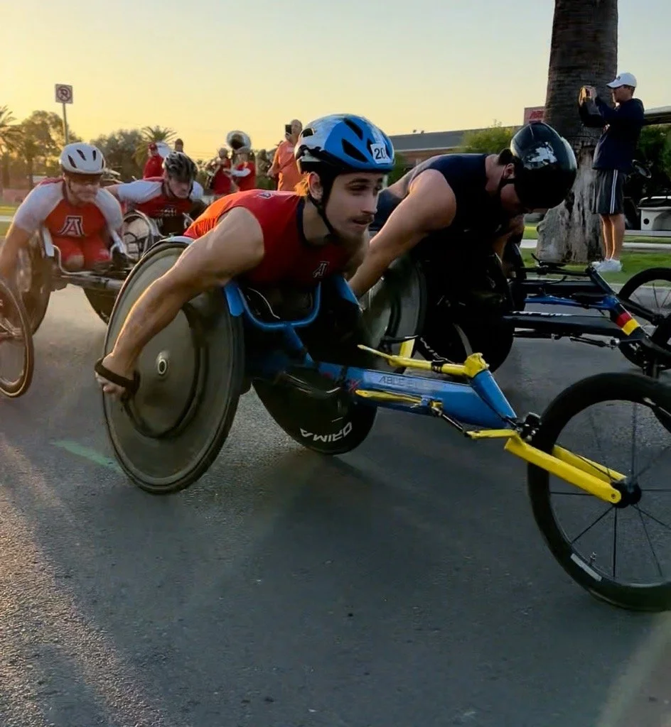 A group of athletes in wheelchairs participating in a race or event, with some people standing and taking pictures in the background during sunset.