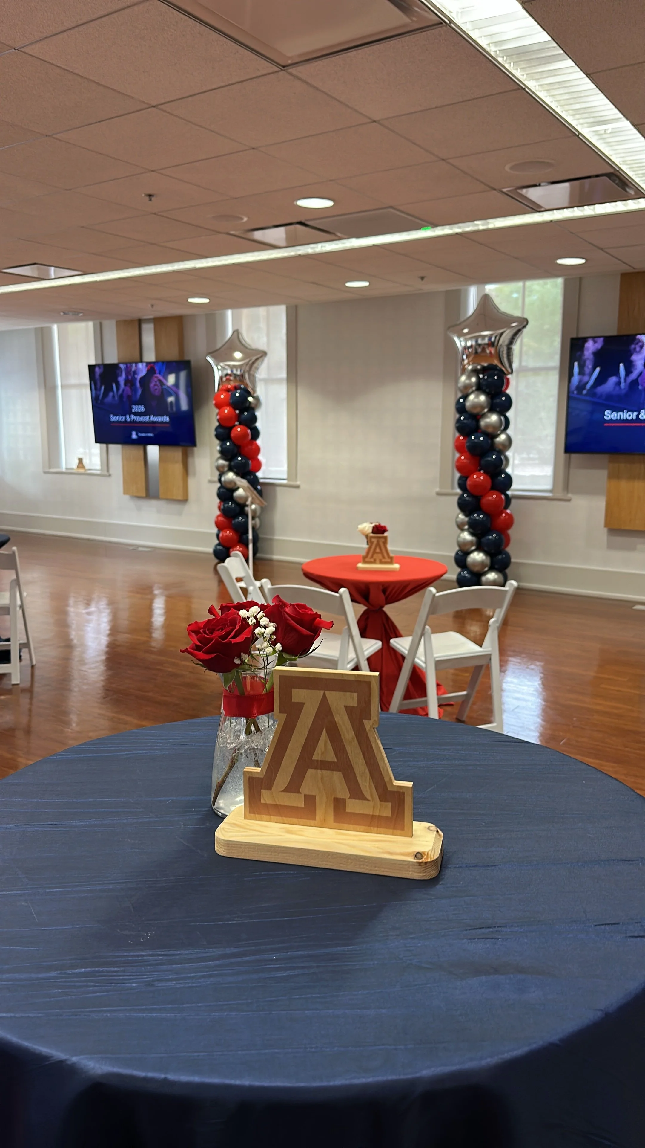 Decorative tables with floral arrangements and university-themed decorations, set up for an awards event in a banquet hall, with balloon columns and large screens in the background.