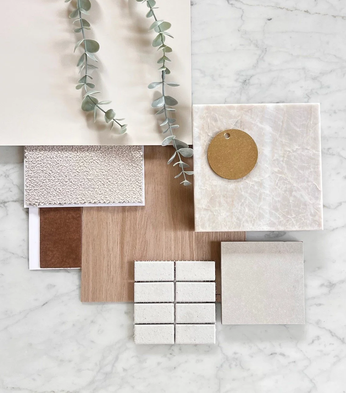 A flatlay of design materials on marble. Features sprigs of eucalyptus, textured samples, a wood plank, tiles, and a circular swatch conveying a calm, natural tone.