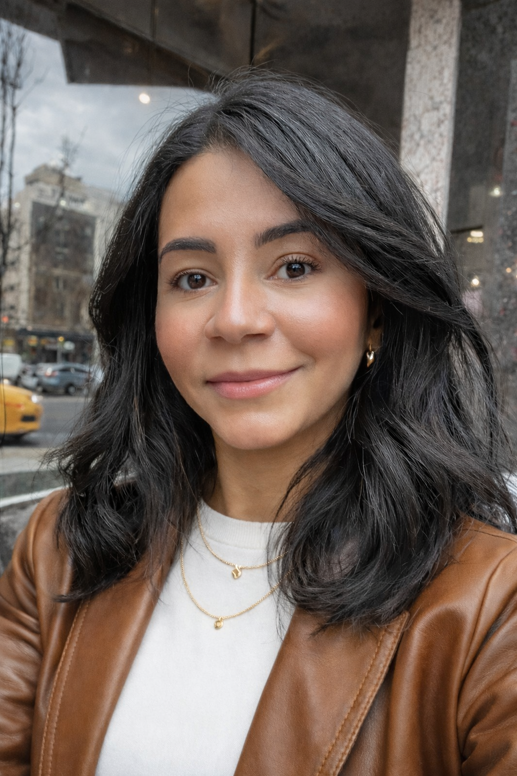 Kercia with shoulder-length dark hair smiles slightly in an urban setting. She wears a brown jacket and necklace. Background includes a blurred street view with cars.