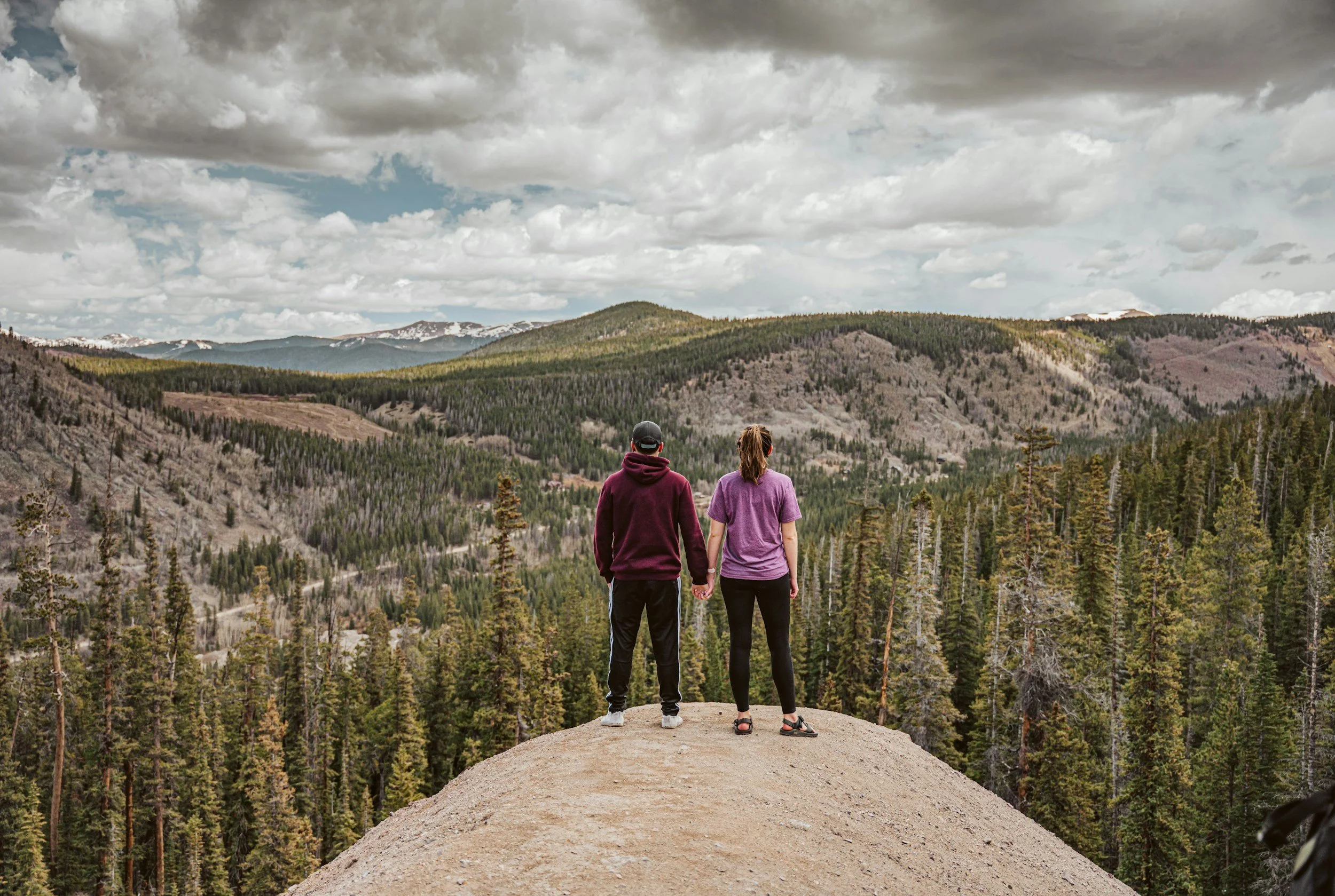 Two people holding hands while standing on a hilltop overlooking a forested valley and mountains under cloudy skies.