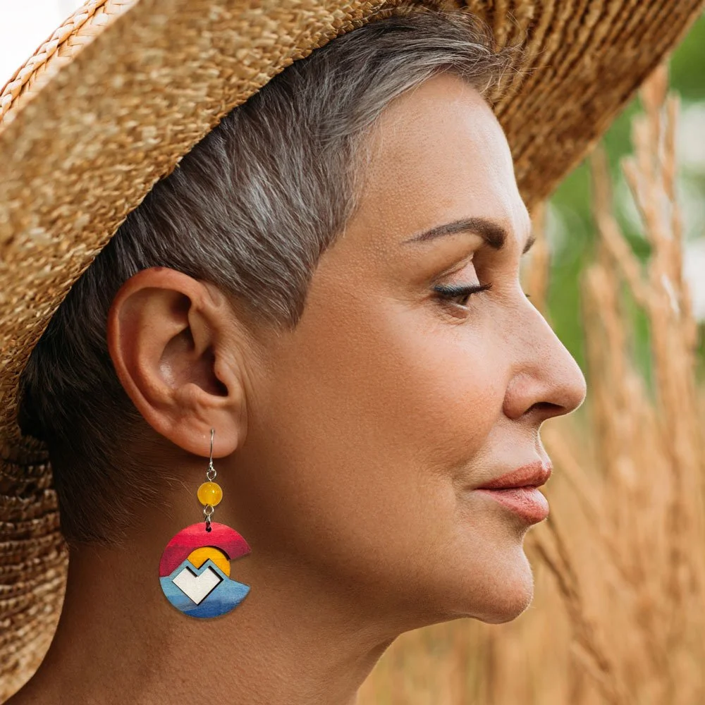 Profile of an older woman with short gray hair wearing a woven sun hat and colorful geometric earrings, standing outdoors near tall grass or plants.