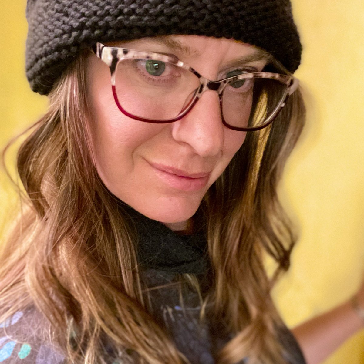 A woman wearing glasses, a black knit hat, and a black scarf, smiling softly at the camera against a yellow background.