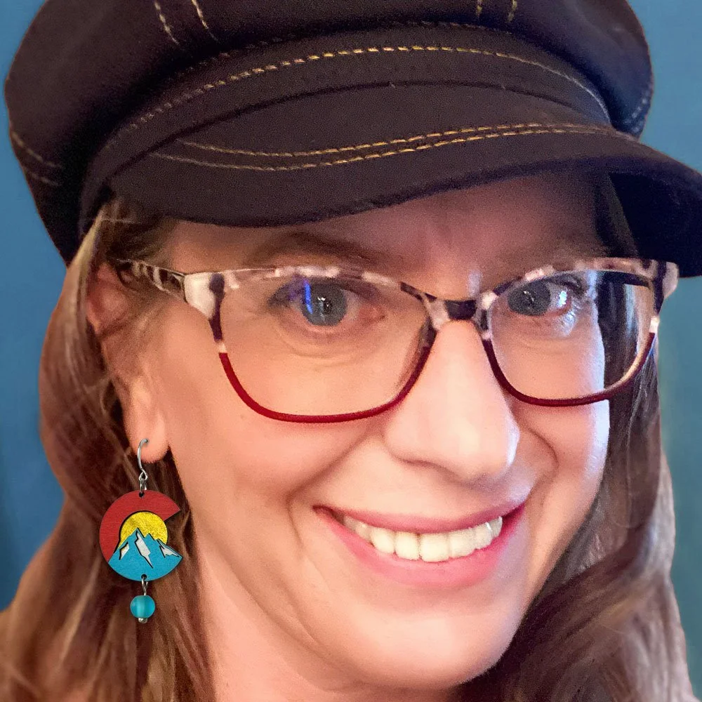 A woman with glasses, a black cap with yellow stitching, and a mountain-themed earring, smiling at the camera against a blue background.