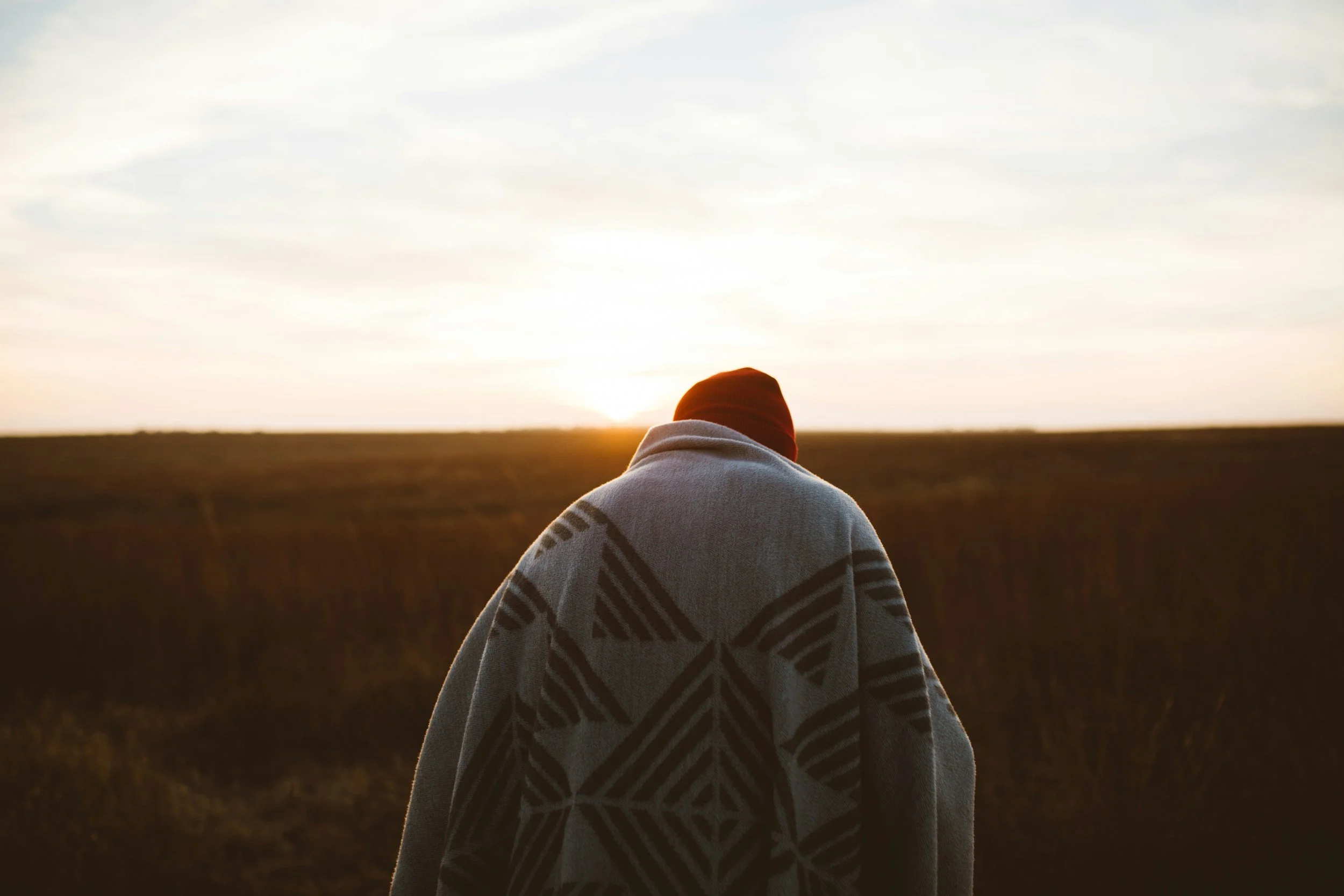 Person wearing a patterned blanket or cloak, standing outdoors in a field at sunset or sunrise.