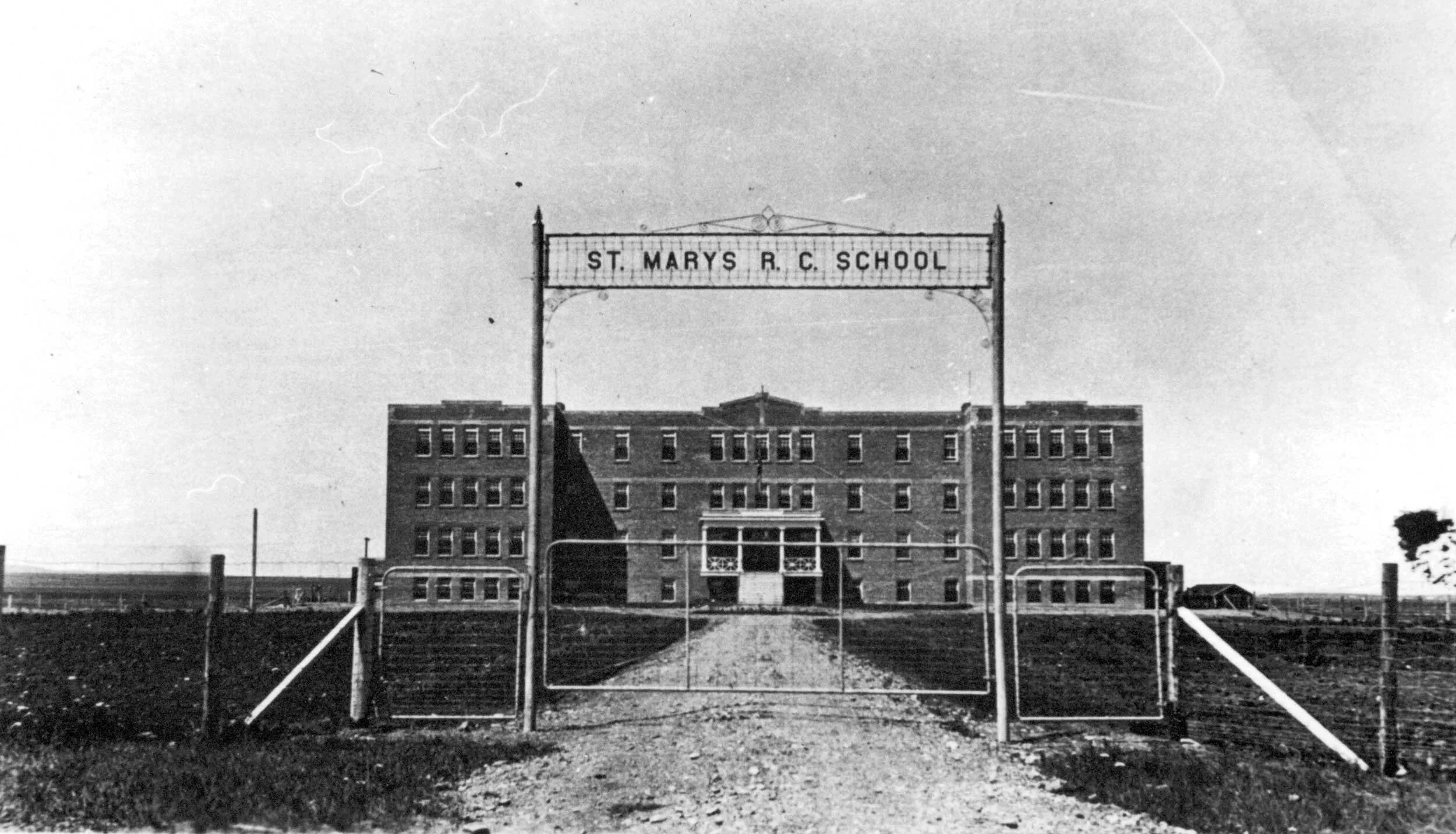 Black and white photo of the front entrance to St. Marys R.C. School, with a large brick building behind a gated fence under a clear sky.