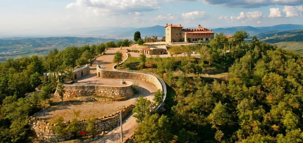 A castle on top of a hill with a winding stone wall pathway surrounded by green trees, overlooking a valley with mountains in the distance under a partly cloudy sky.