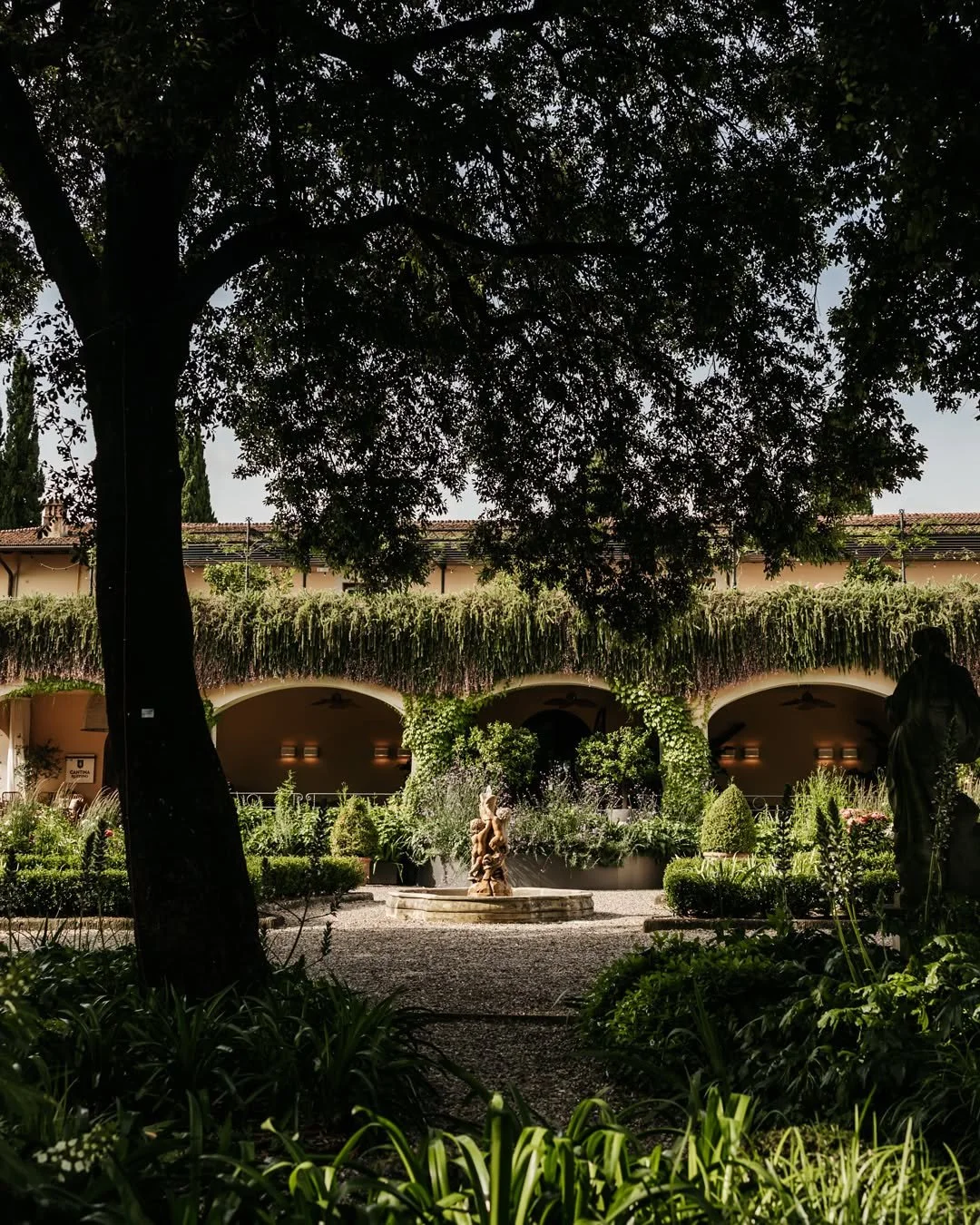 A courtyard with lush green plants and a fountain at the center, surrounded by a building with arches and greenery cascading from the roof, large trees providing shade.