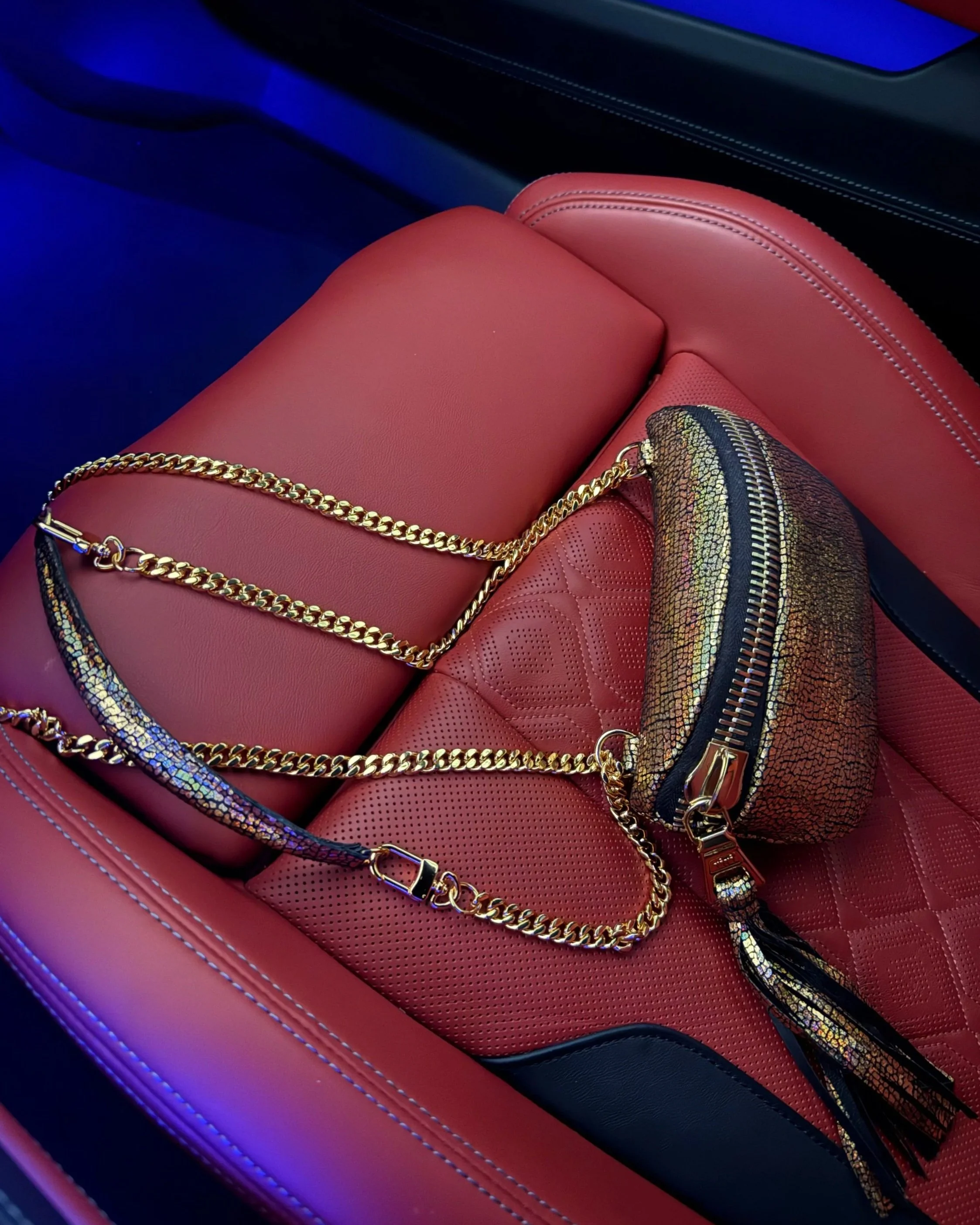 Gold iridescent leather handbag with a chain strap styled on a red leather car seat.