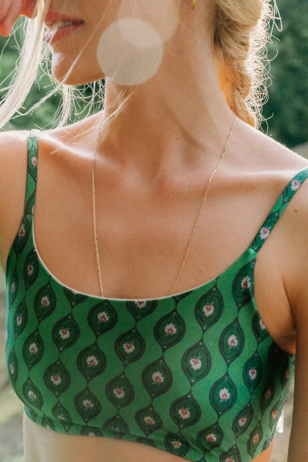 Mulher vestindo top verde com padrão floral e uma corrente dourada, ao ar livre com fundo de árvores.