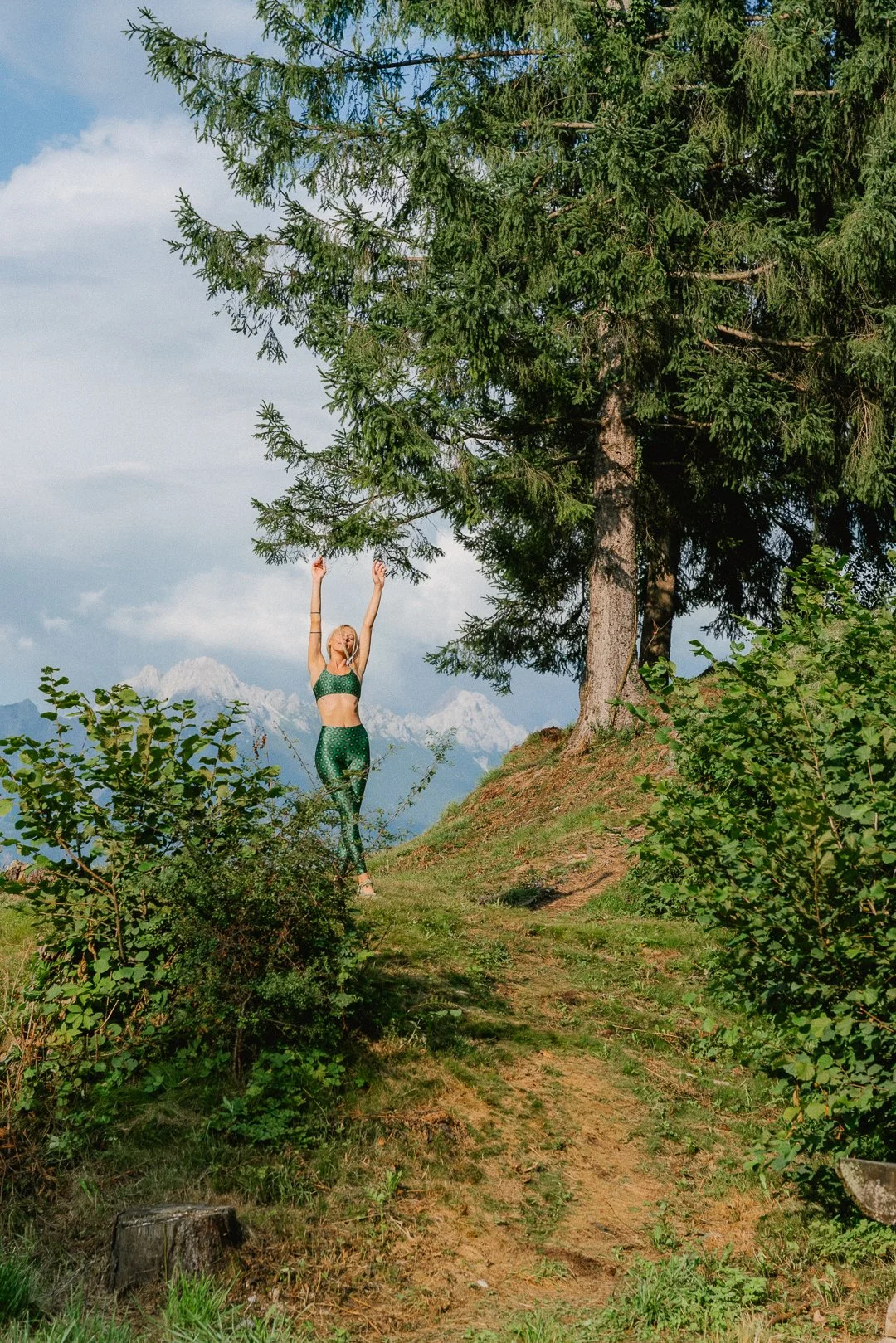 Mulher praticando ioga ao ar livre em uma trilha na floresta com árvores verdes e montanhas ao fundo.