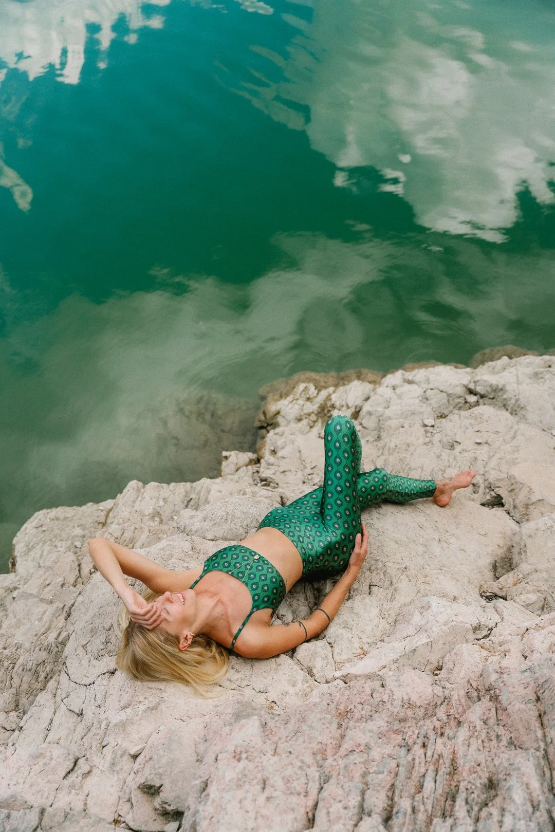 Mulher deitada sobre rochas ao lado de uma água verde, usando roupa de banho verde com padrão circular, com cabelo loiro, cabeça apoiada na mão, sorriso, ambiente natural.