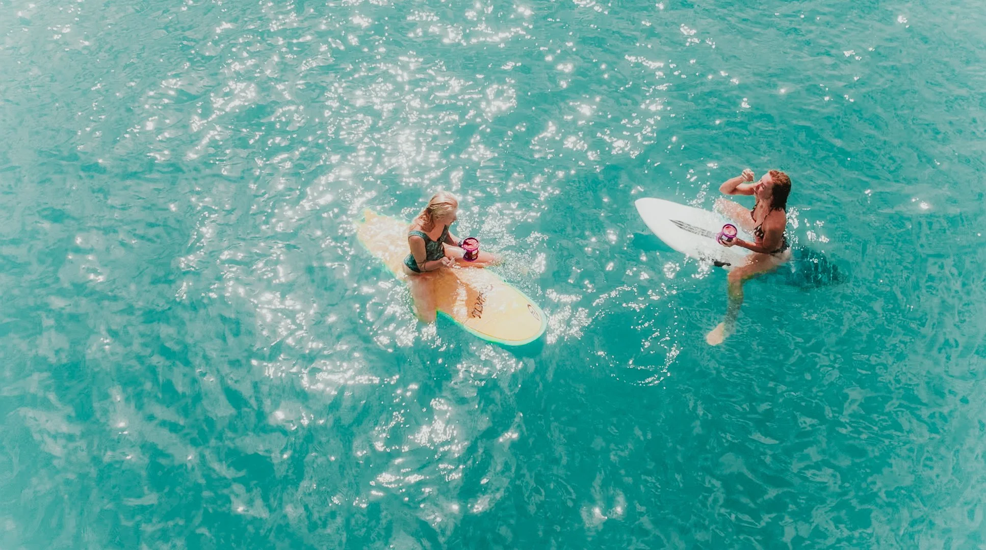 Duas mulheres surfando na água, usando pranchas de surf, uma delas segurando uma garrafa de água rosa.