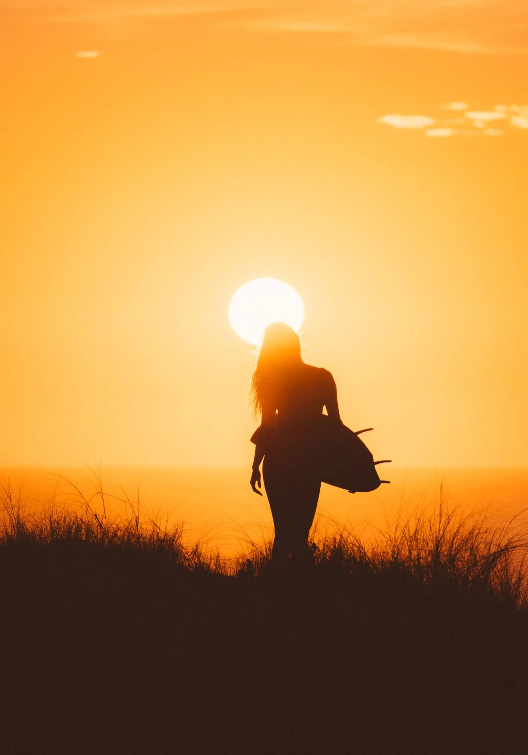 Silhueta de uma mulher carregando uma prancha, caminhando na areia ao pôr do sol com céu laranja.