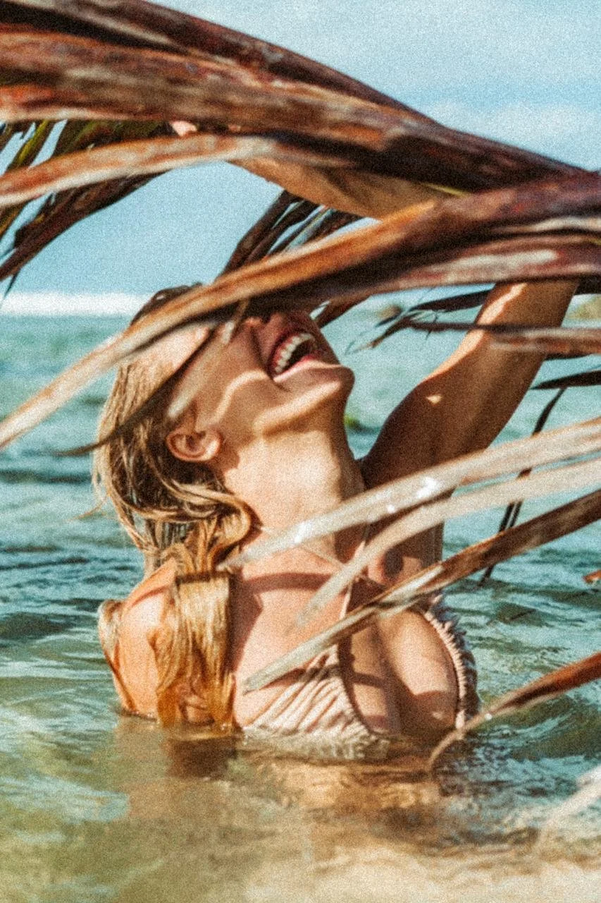 Mulher sorrindo na água do mar, parcialmente escondida por folhas secas de palha.