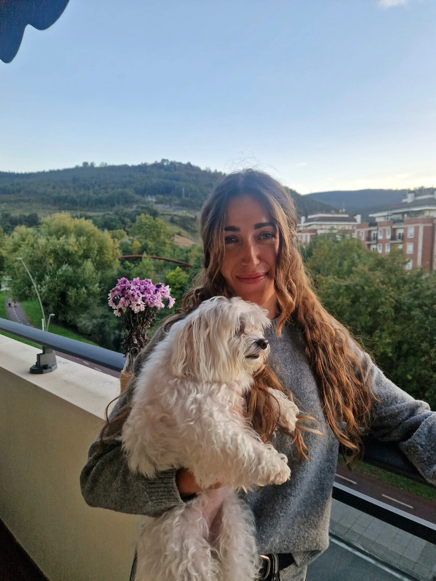 Mujer sonriendo sosteniendo un perro blanco de pelo rizado en un balcón con vista a áreas verdes y colinas en el fondo.