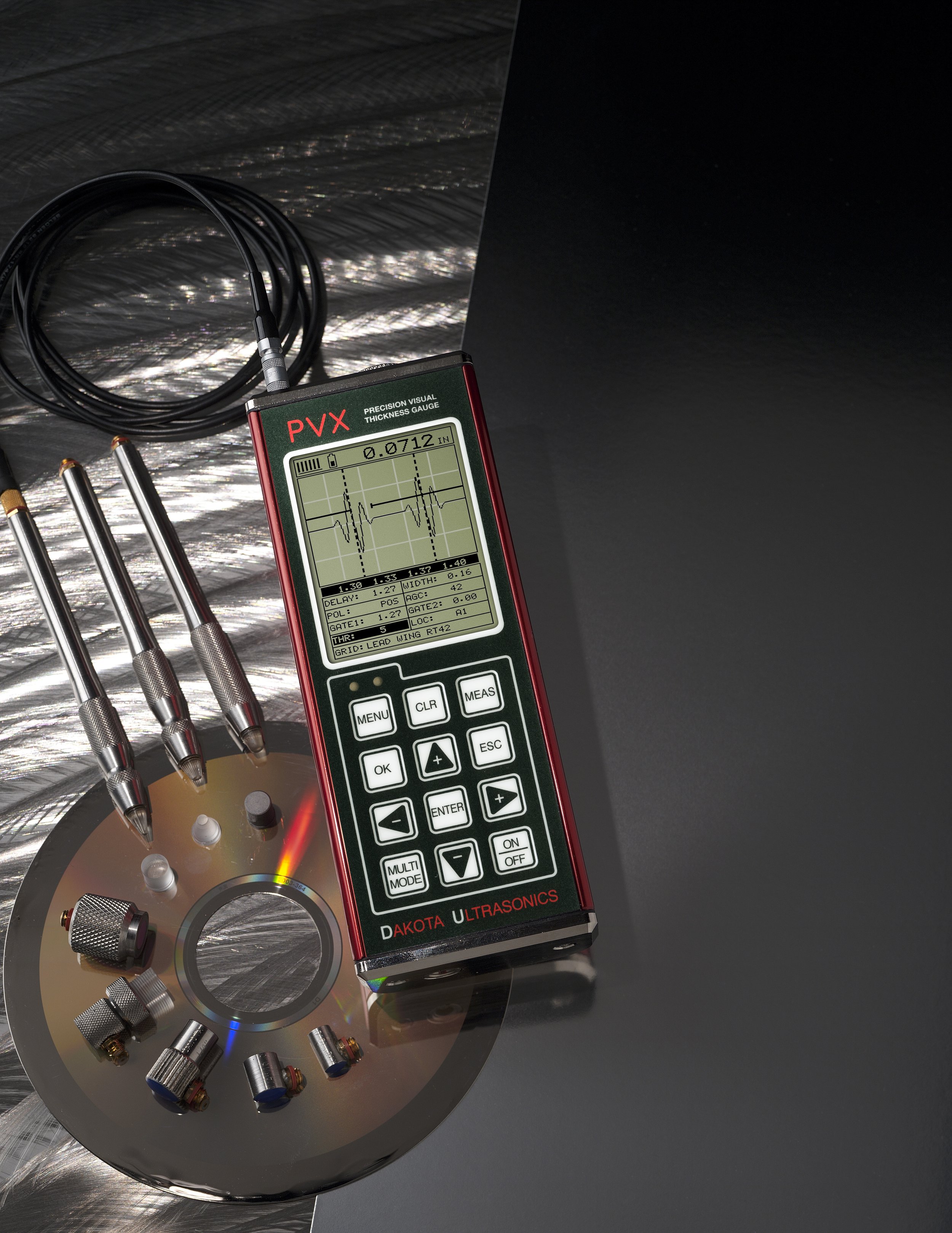 Instrumento de medición llamado dureza o espesor con pantalla digital y cables conectados, junto a un disco de metal con pequeños componentes de metal en su superficie, y varias pequeñas herramientas metálicas con diferentes puntas calibradas sobre una superficie metálica.