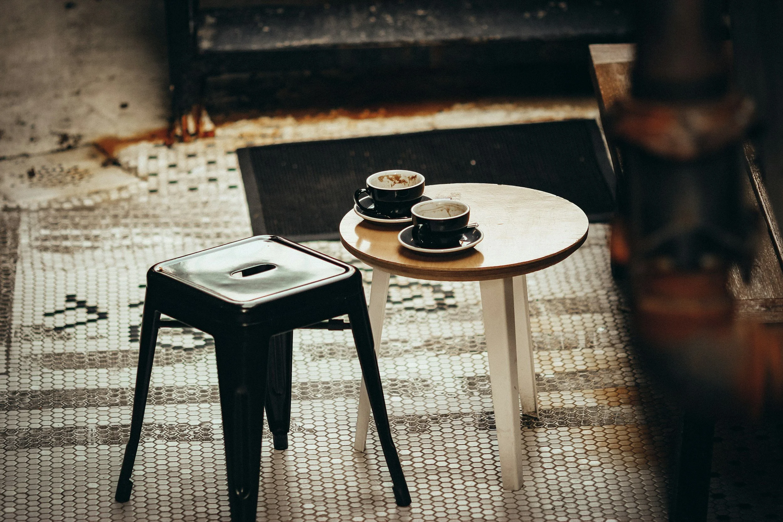 Two black coffee cups on saucers and a half-empty cup on a small round table in a cozy cafe with black and white tiled floor.