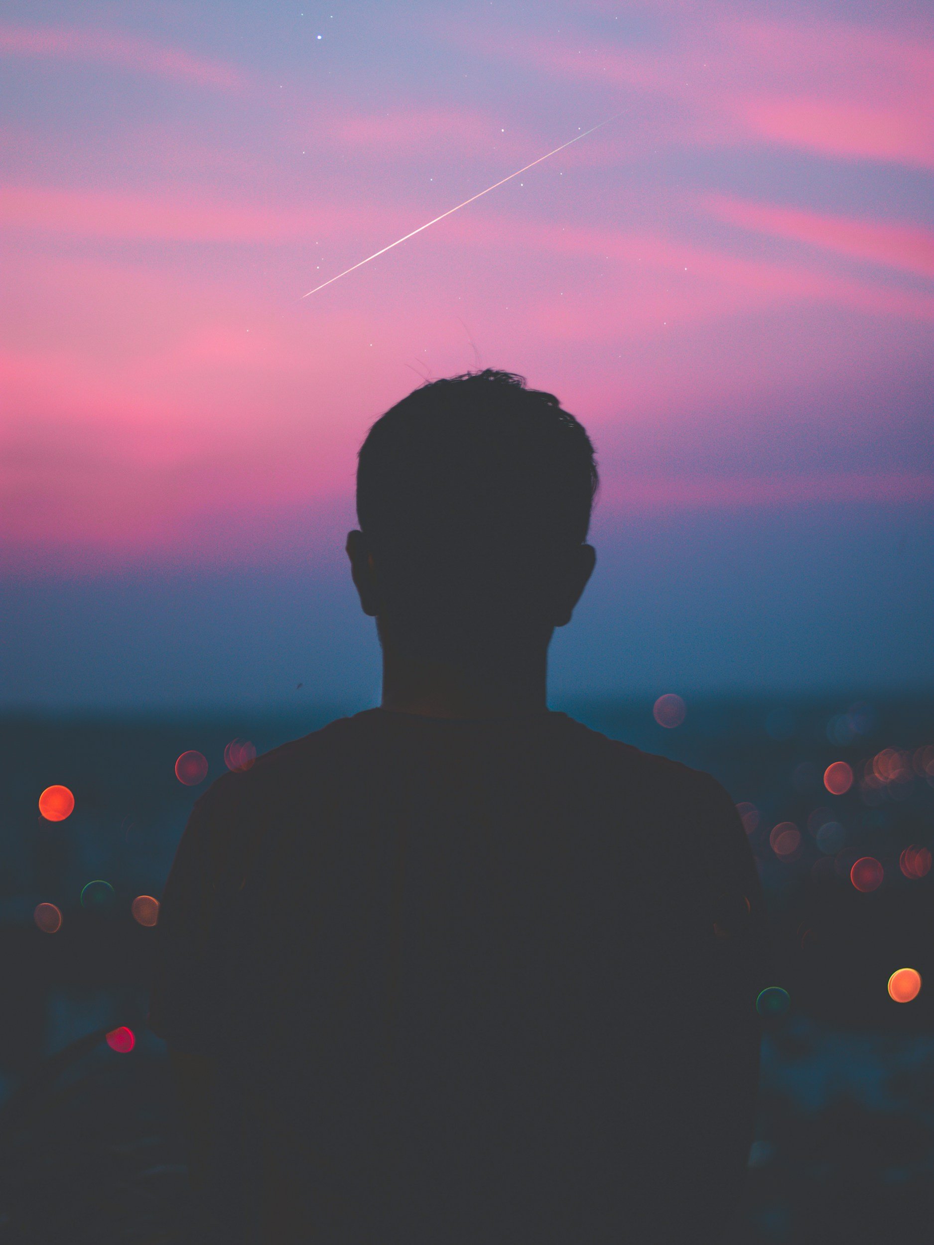 Silhouette of a man watching the sky at dusk with pink and purple clouds, stars, and a shooting star.