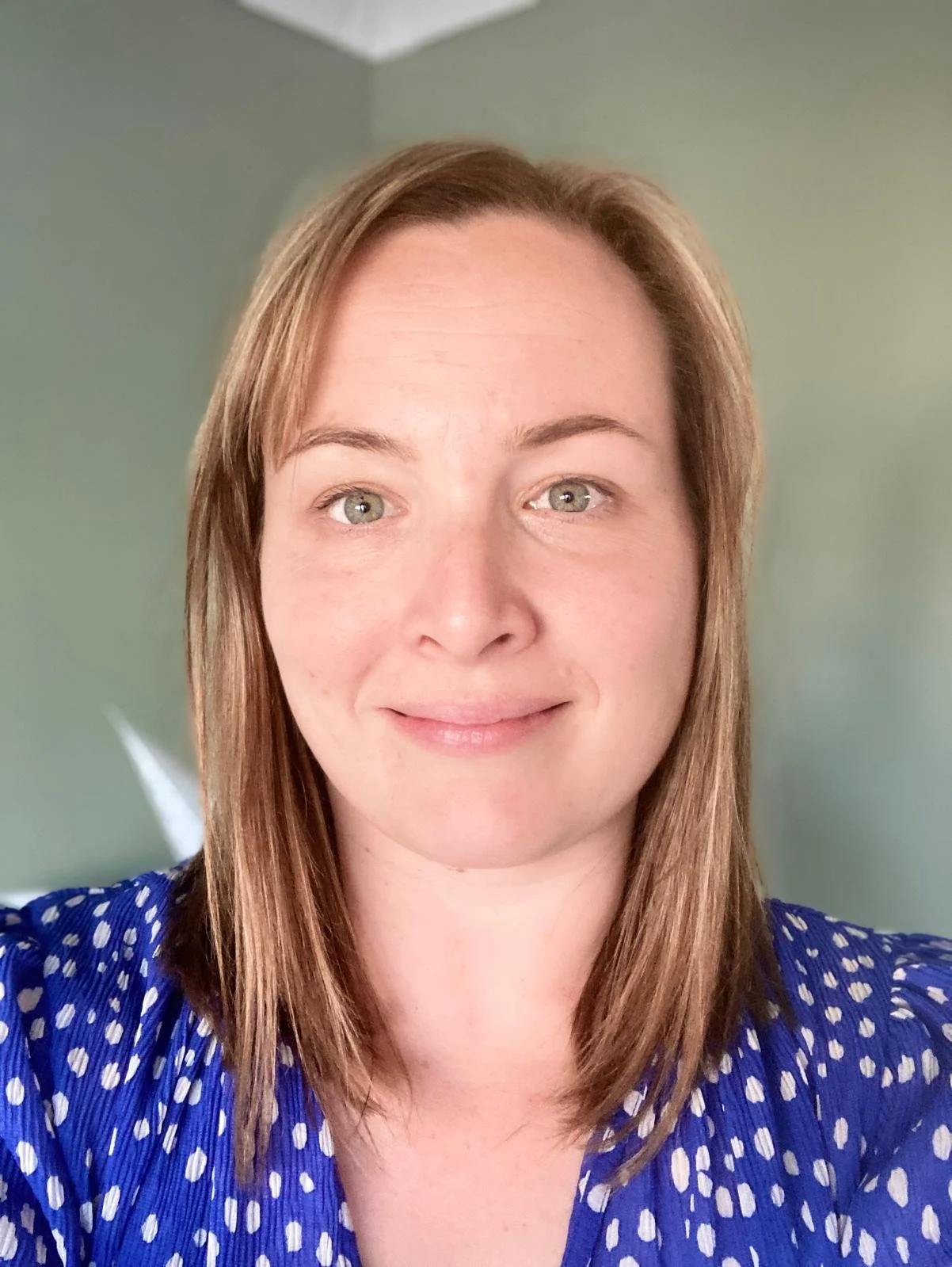 Close-up of a woman with shoulder-length brown hair and green eyes, smiling, wearing a blue polka-dot shirt with a light green wall in the background.