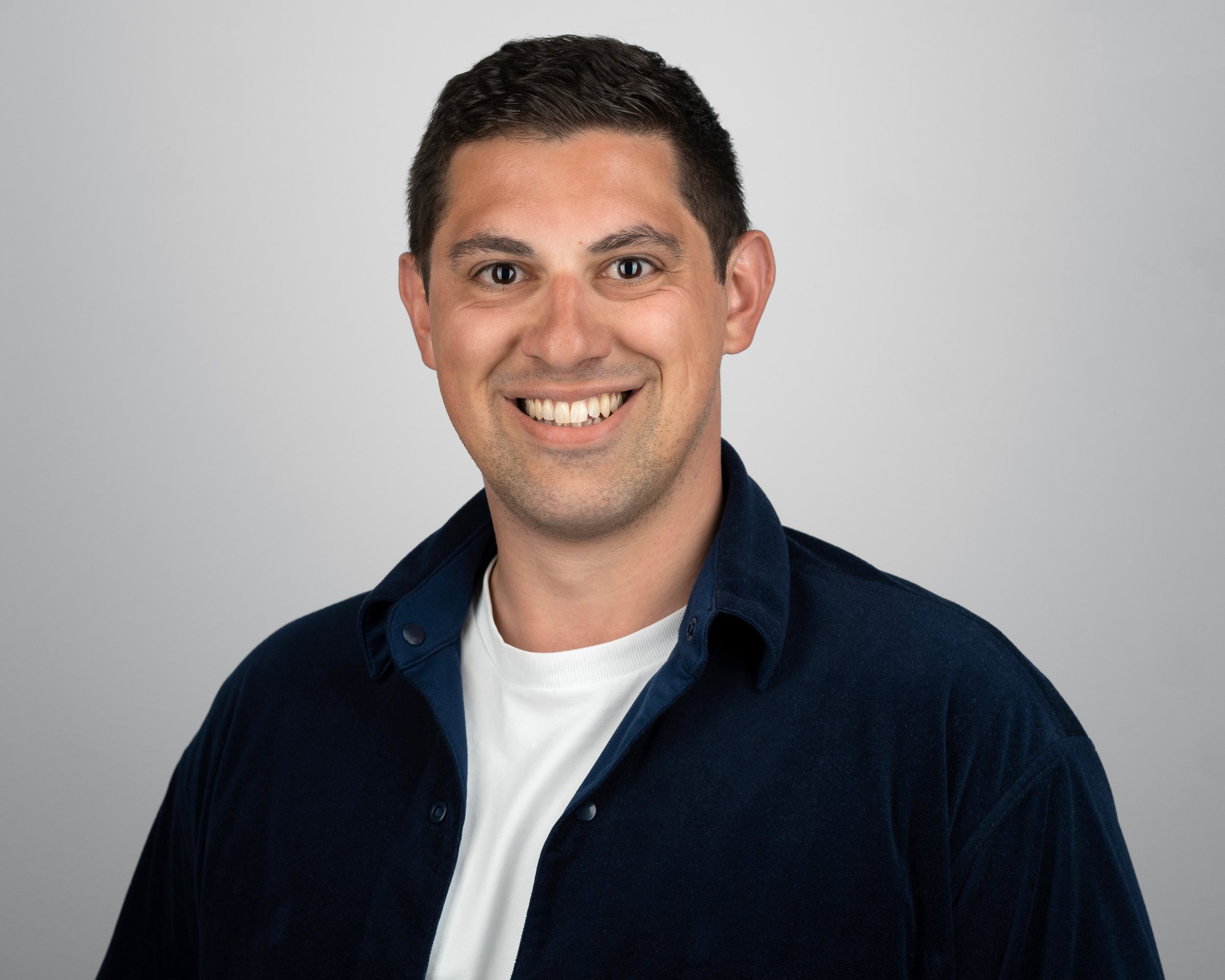 A smiling man with short dark hair wearing a navy blue shirt and white t-shirt against a gray background.