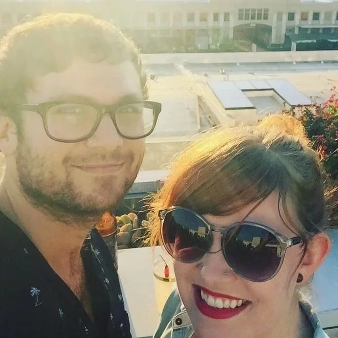 A man and woman smiling on a rooftop during sunset, with city buildings in the background. The woman is wearing large sunglasses and has red lipstick, while the man is wearing glasses and has a beard.