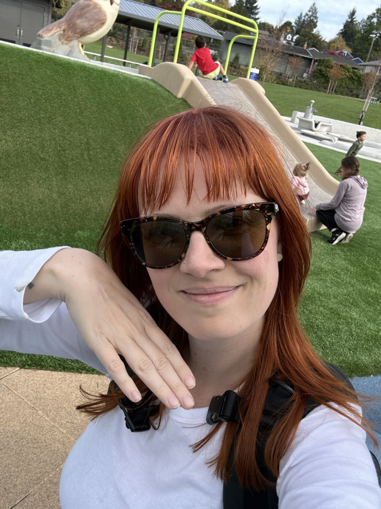 A woman with red hair, wearing sunglasses and a white shirt, taking a selfie at a playground. In the background, children and adults are playing on a slide and other playground equipment, with green grass and trees around.