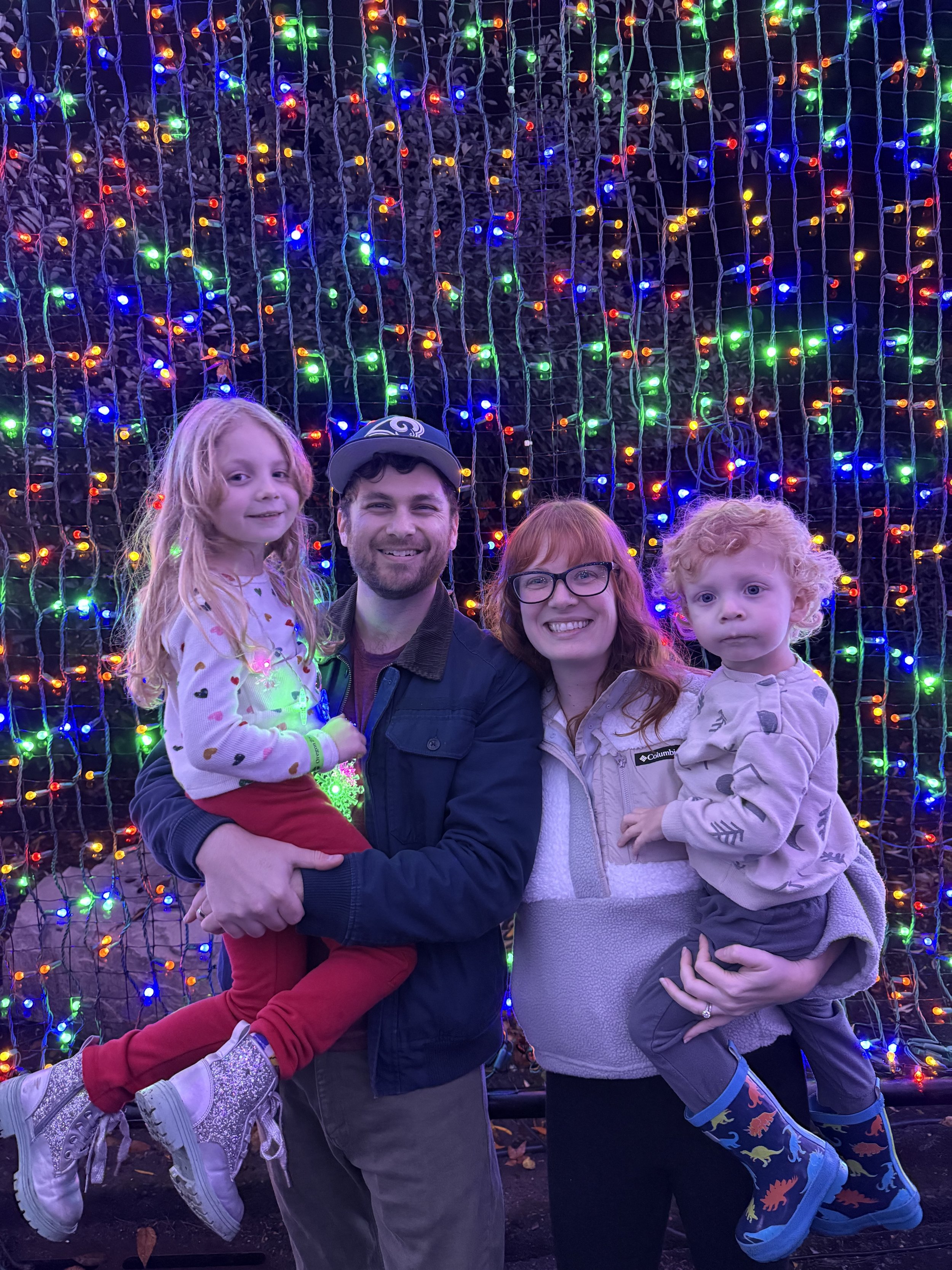 Family of four standing in front of a colorful LED holiday light display, with the father holding a young girl and the mother holding a young boy, all smiling.