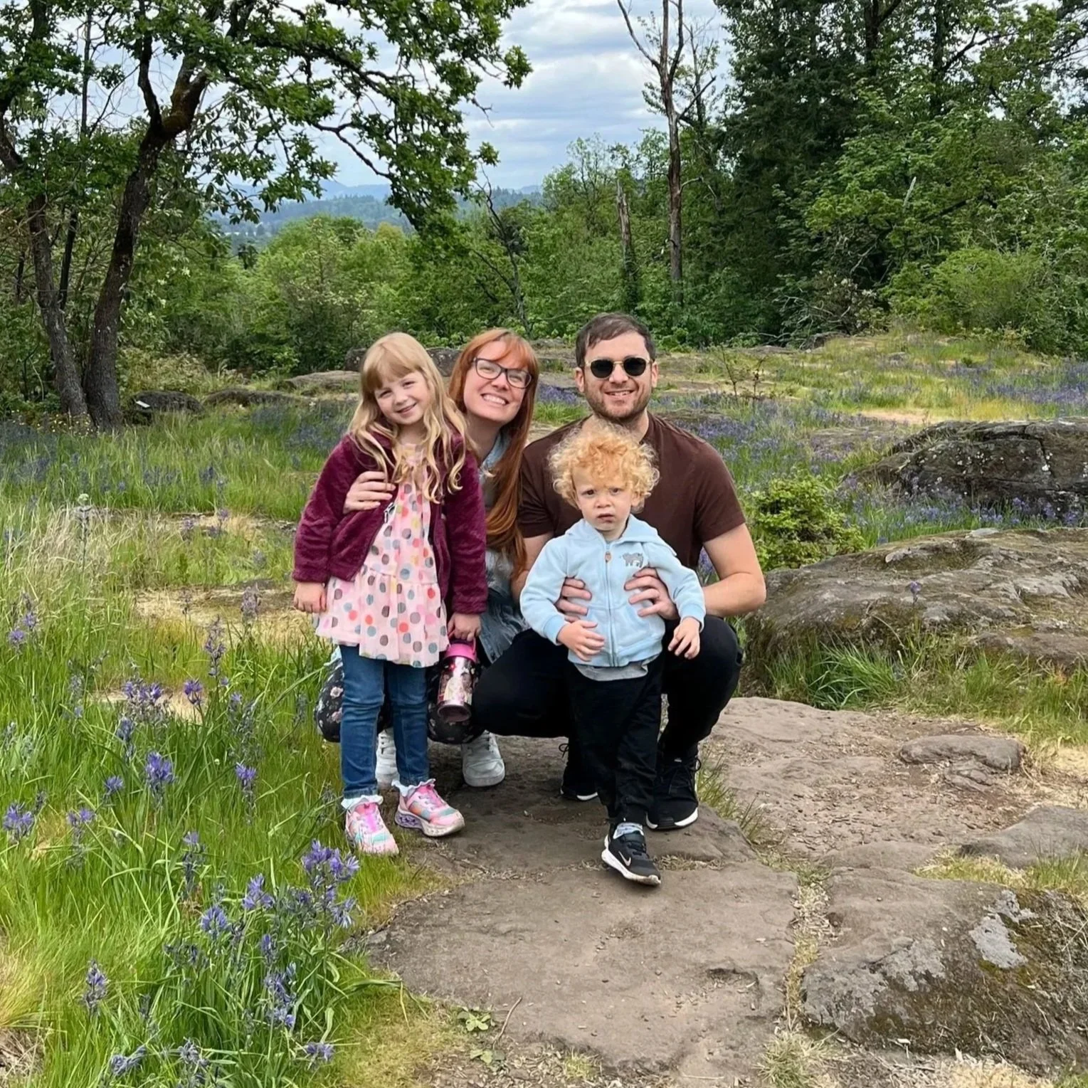 A family of four posing outdoors on a trail in a green, forested area with trees, rocks, and purple wildflowers.