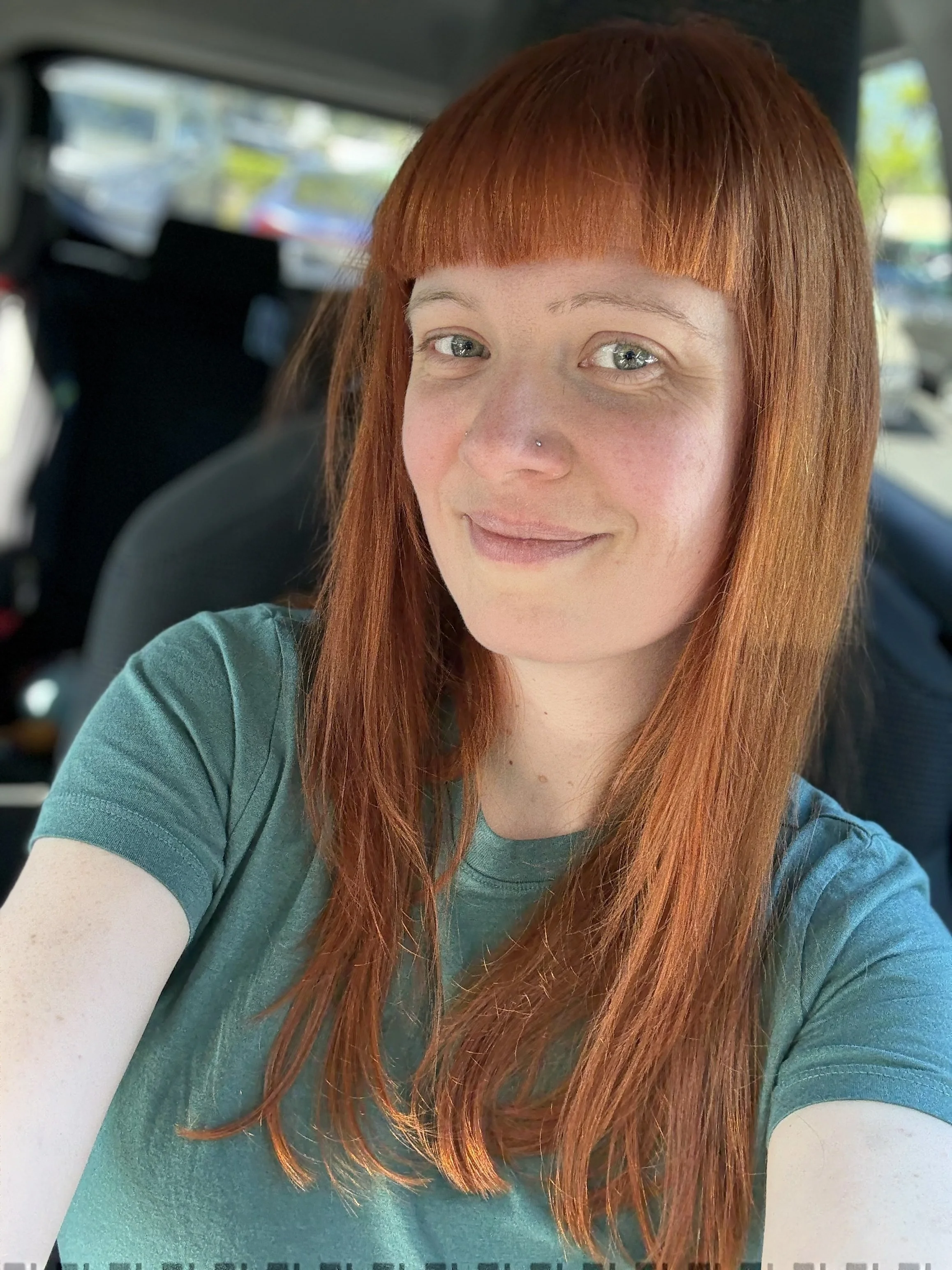 A woman with long red hair and a nose piercing taking a selfie inside a vehicle, with other vehicles and trees visible outside the window.