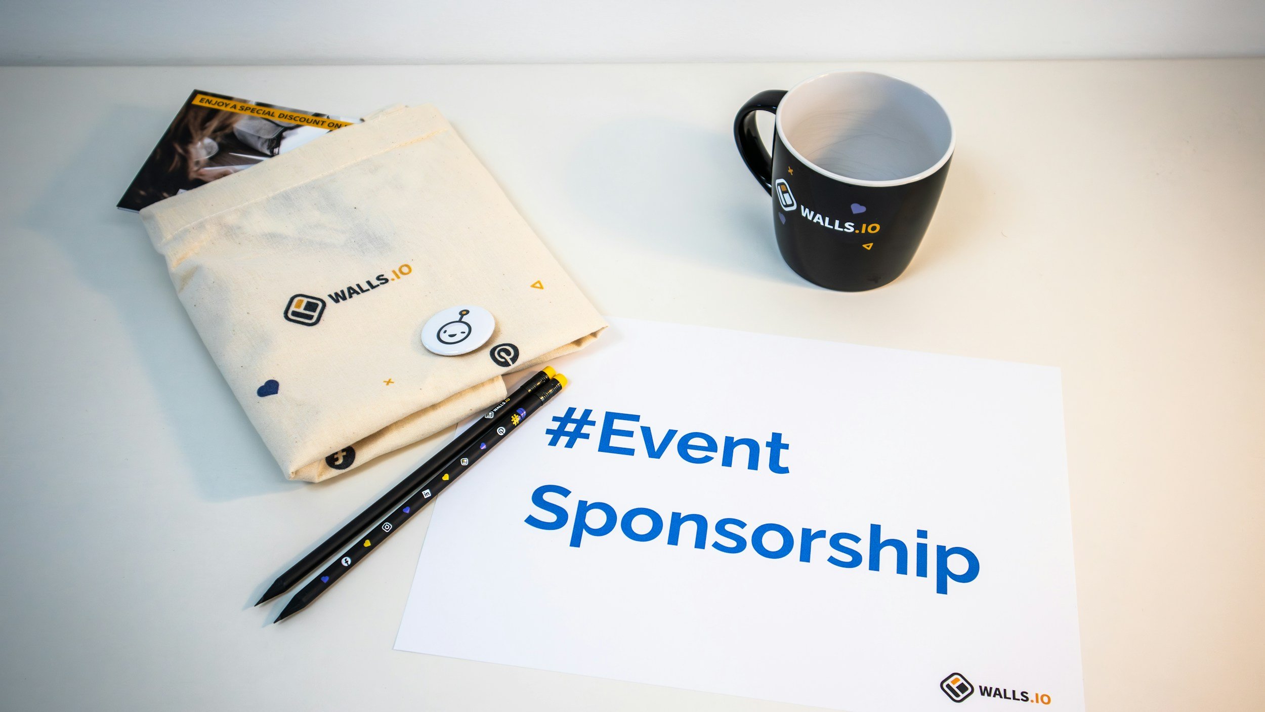 A black coffee cup sitting atop a set of pencils and a tote bag with the hashtag Event Sponsorship in blue beneath