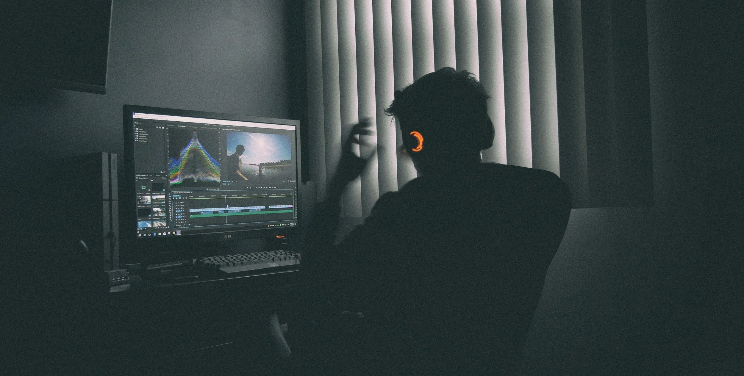 Person working on video editing or color grading on a desktop computer in a dimly lit room, with vertical blinds on the window behind.